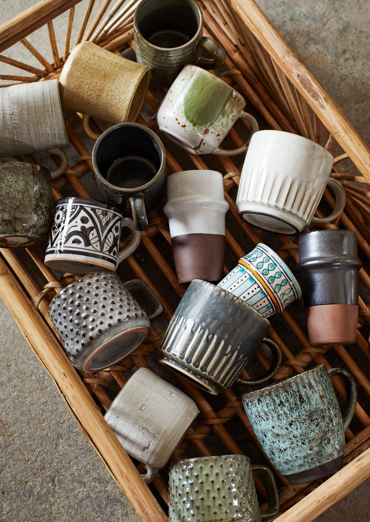 A collection of various textured ceramic mugs by Madam Stoltz arranged inside a woven rattan basket.