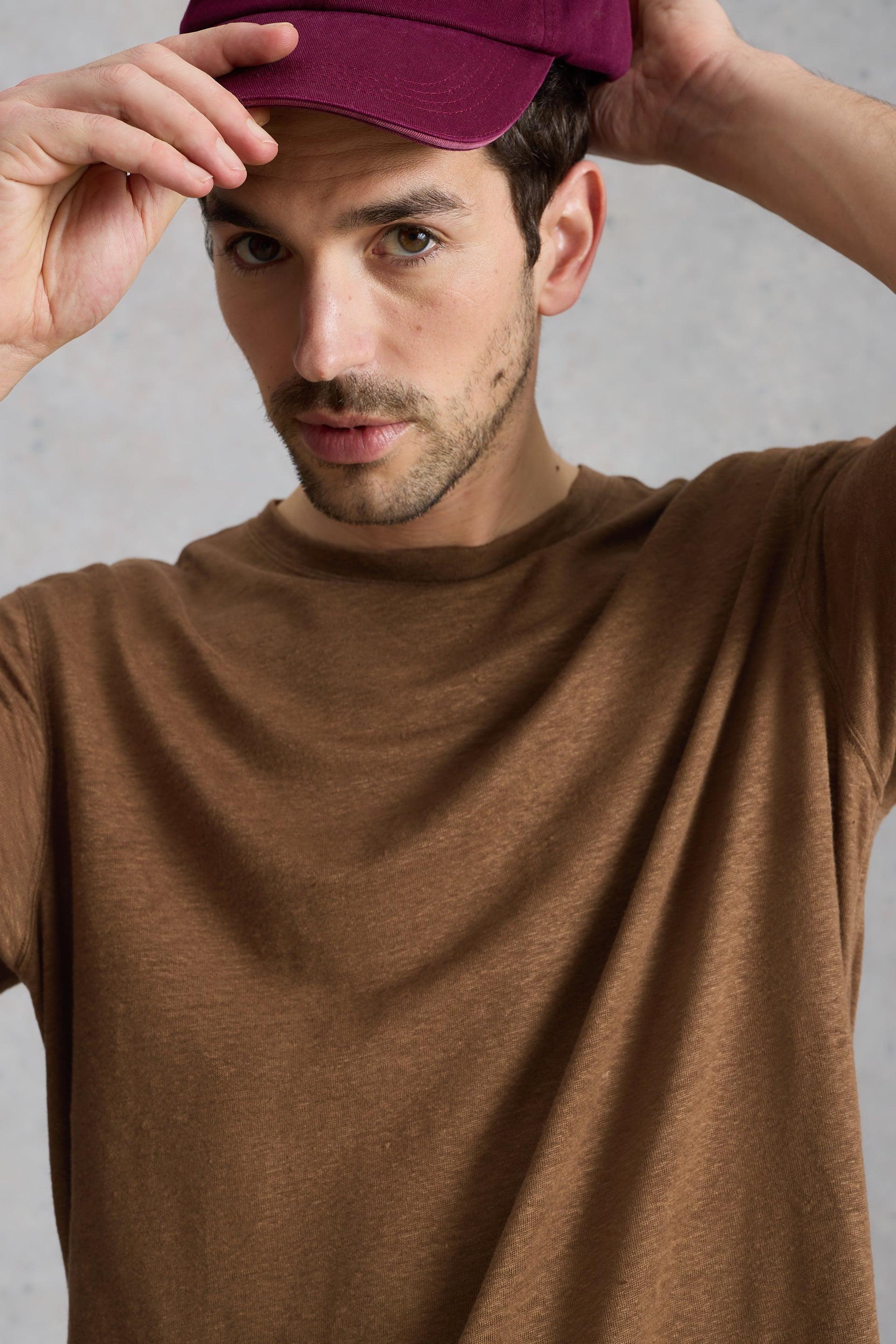 Man wearing a brown sustainable mens linen t-shirt and a maroon baseball cap against a grey backdrop.