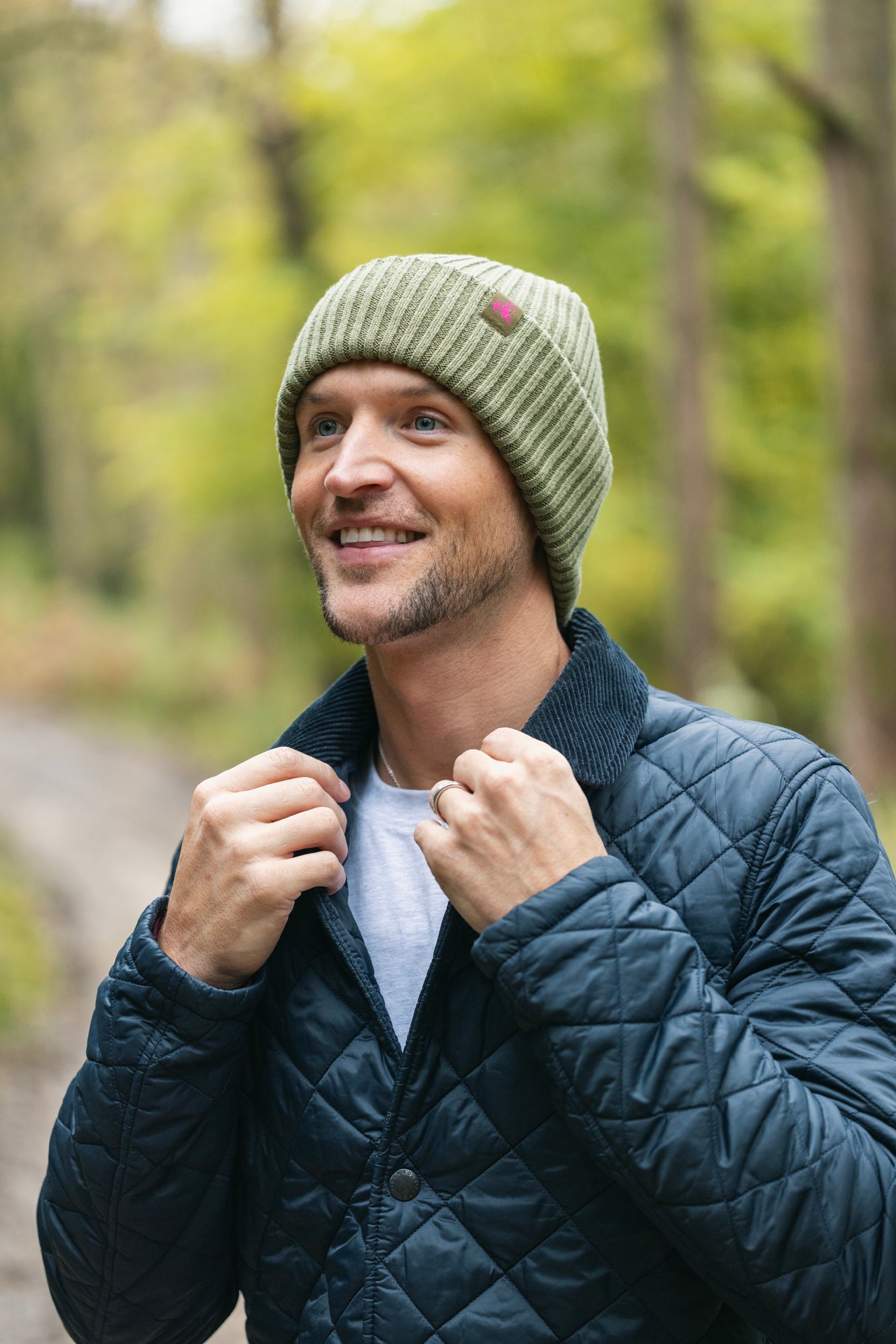 Man wearing a green organic cotton beanie and dark quilted jacket outdoors with trees in the background