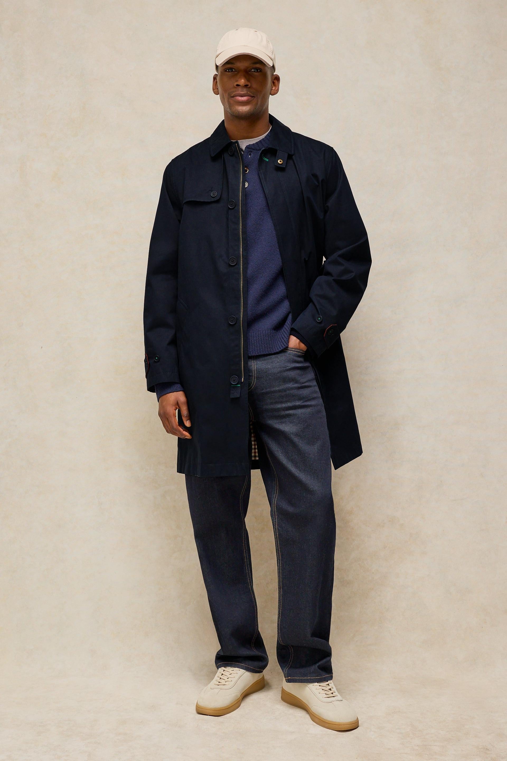 A man modeling a dark navy blue mid-length utility coat over a navy sweater, dark gray trousers, and cream-colored sneakers, standing against a textured beige studio backdrop.