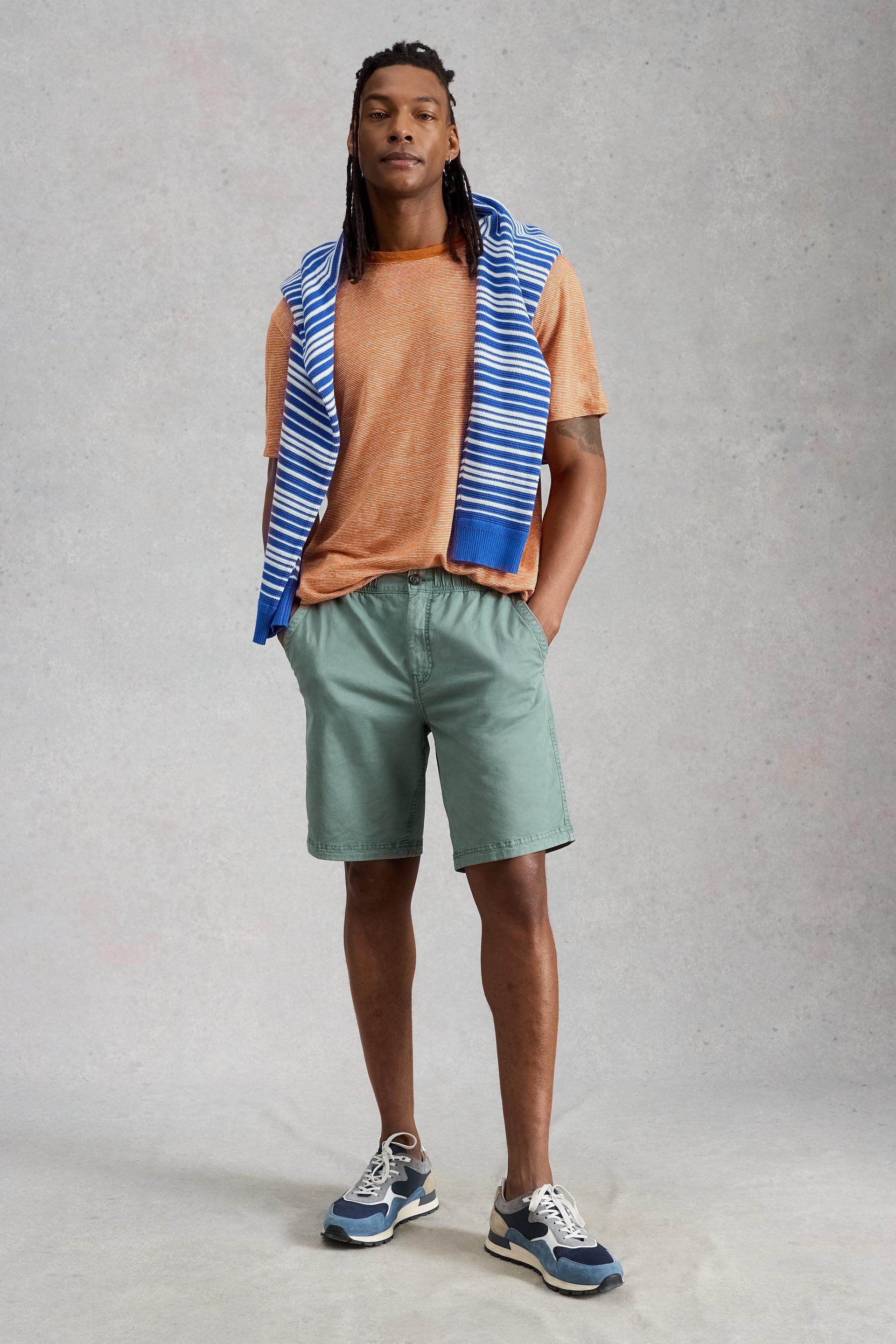 A man wearing an orange 100% linen t-shirt with a blue striped sweater over his shoulders and green shorts.