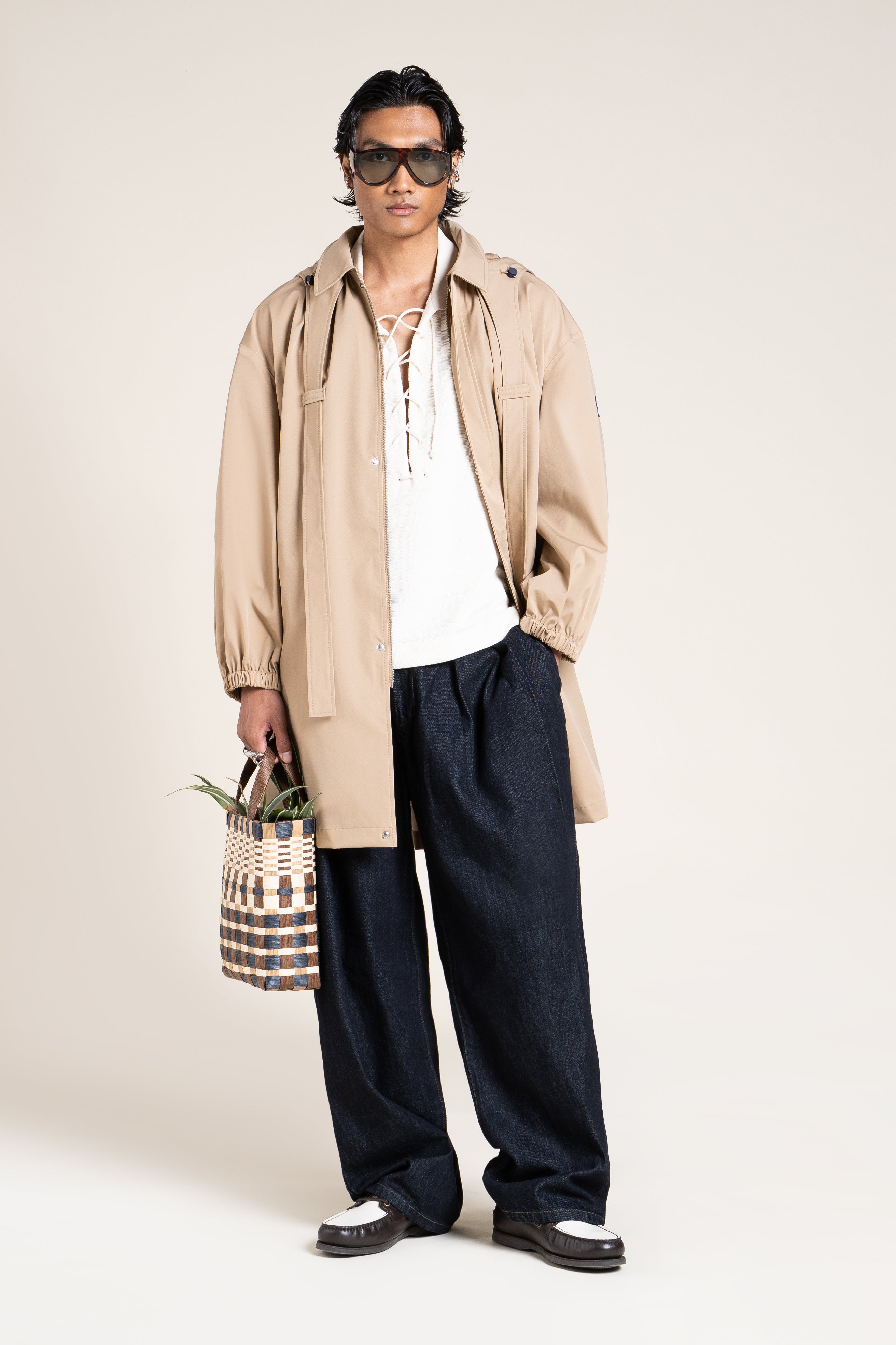 A man wearing a stylish beige waterproof Flotte coat and dark trousers while holding a woven bag against a neutral backdrop.