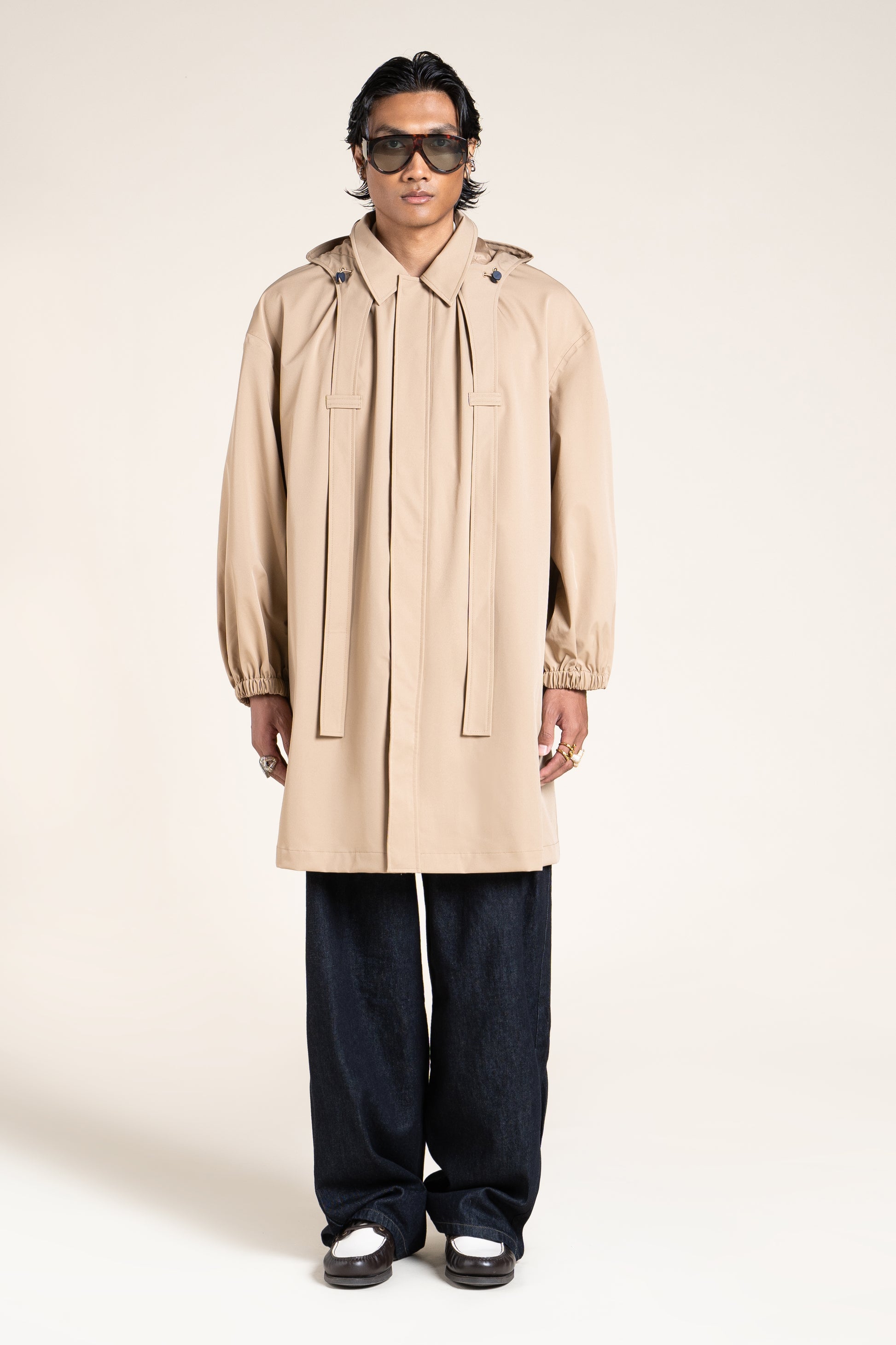 A person standing in a studio wearing a stylish beige waterproof coat with sunglasses