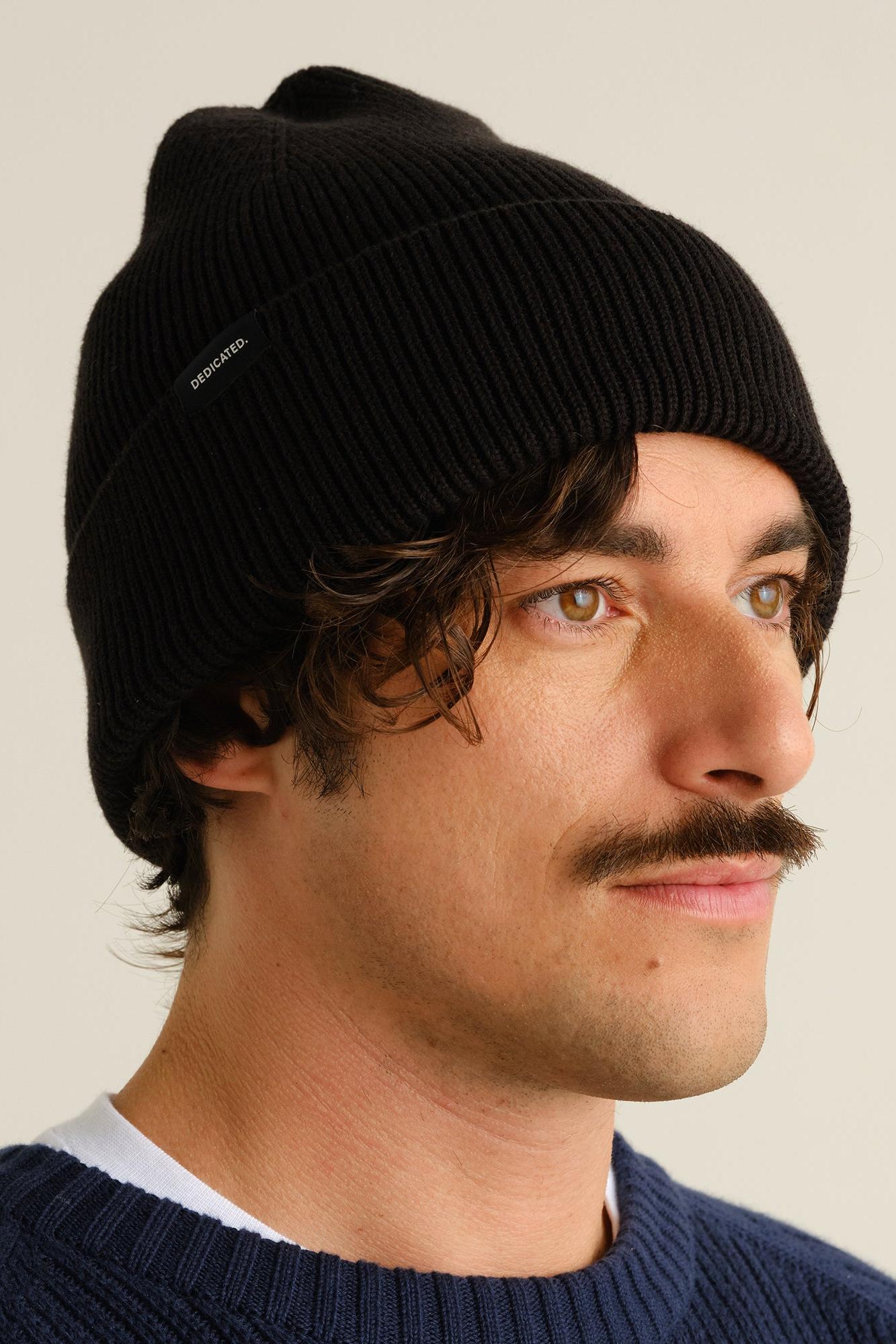 A man wearing a black ribbed knit beanie hat, shown in profile against a plain light beige background.