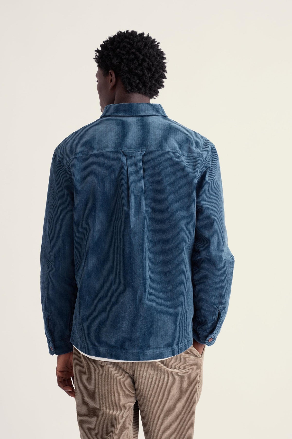 A man seen from behind wearing a blue Seasalt cord casual shirt with tan trousers against a plain light background.