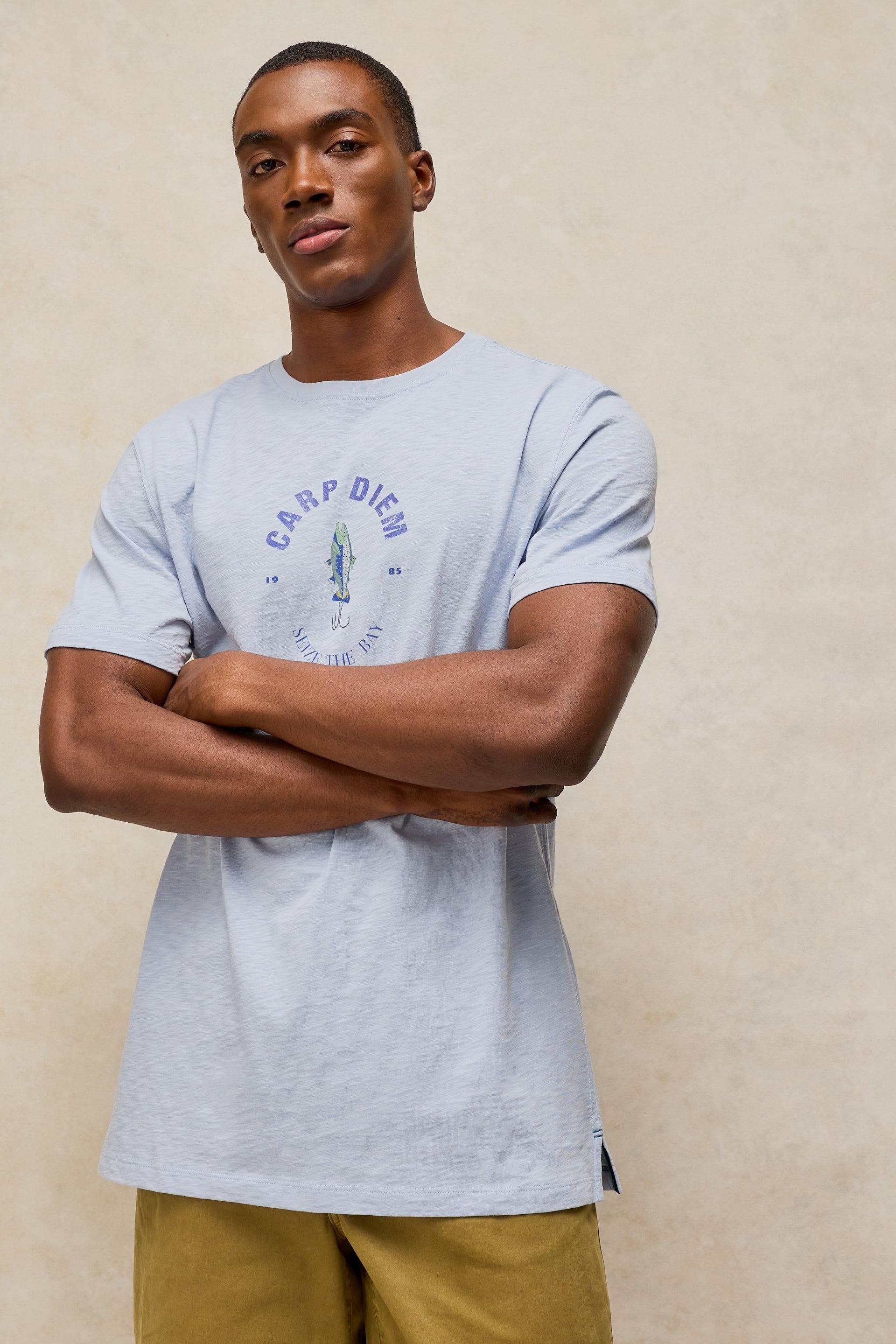 A man wearing a light blue cotton t-shirt with a 'Carp Diem' graphic on the chest, paired with olive green pants, standing against a textured beige wall.