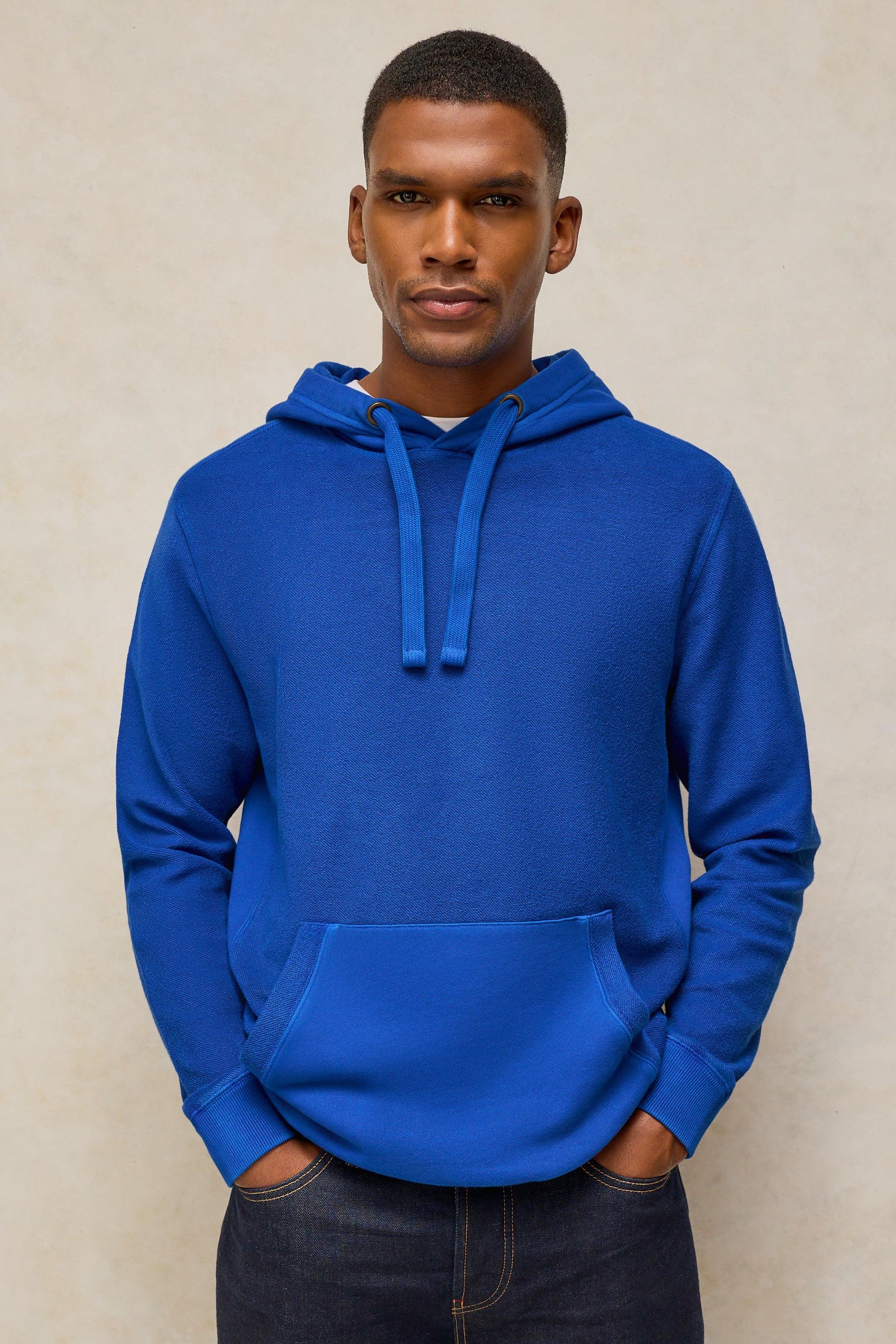 A man wearing a bright royal blue pullover hoodie with matching drawstrings and a front kangaroo pocket, paired with dark jeans, standing against a neutral beige textured background.