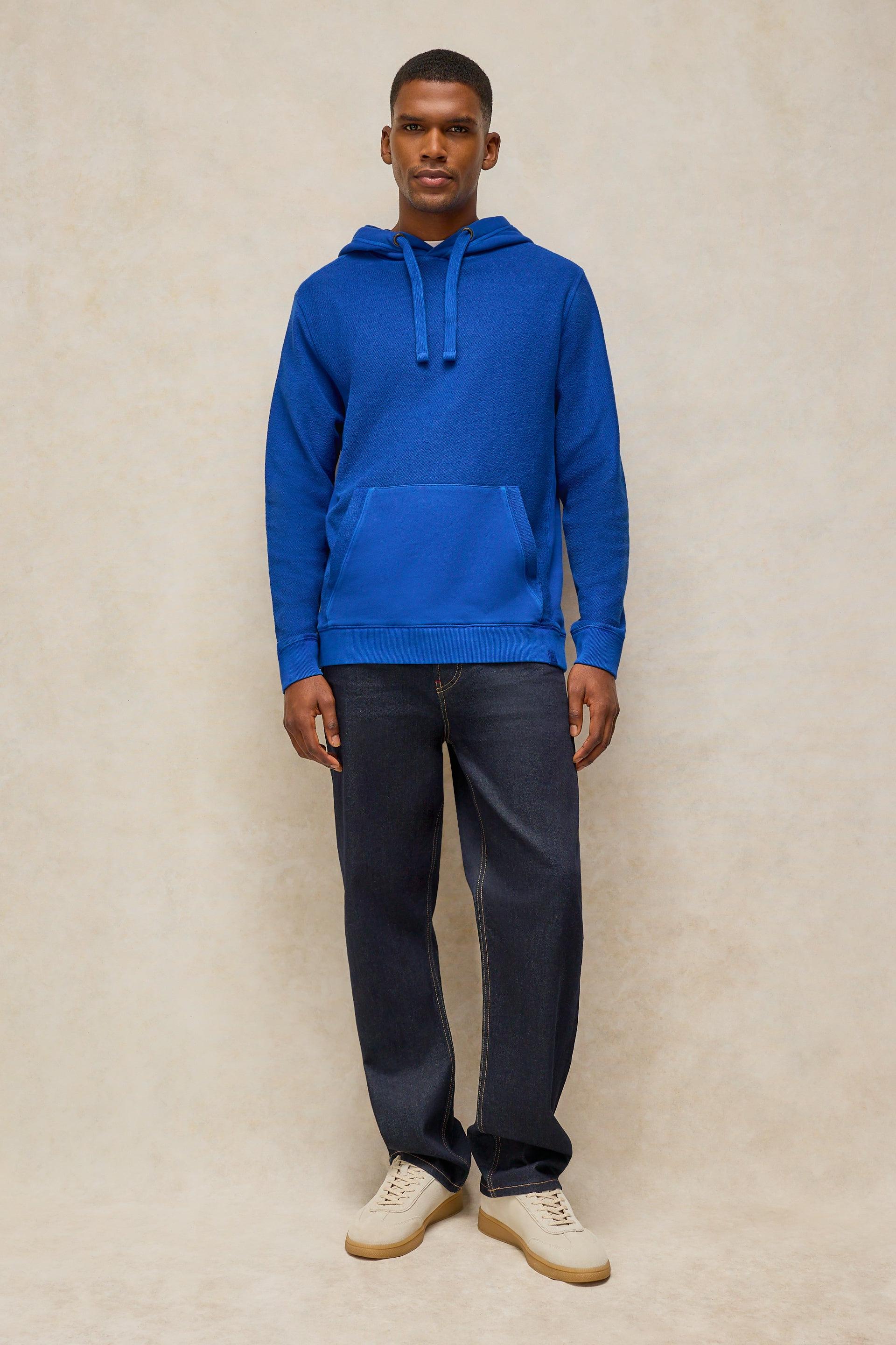 A man wearing a bright blue pullover hoodie with dark blue jeans and beige sneakers, standing against a textured beige studio backdrop.