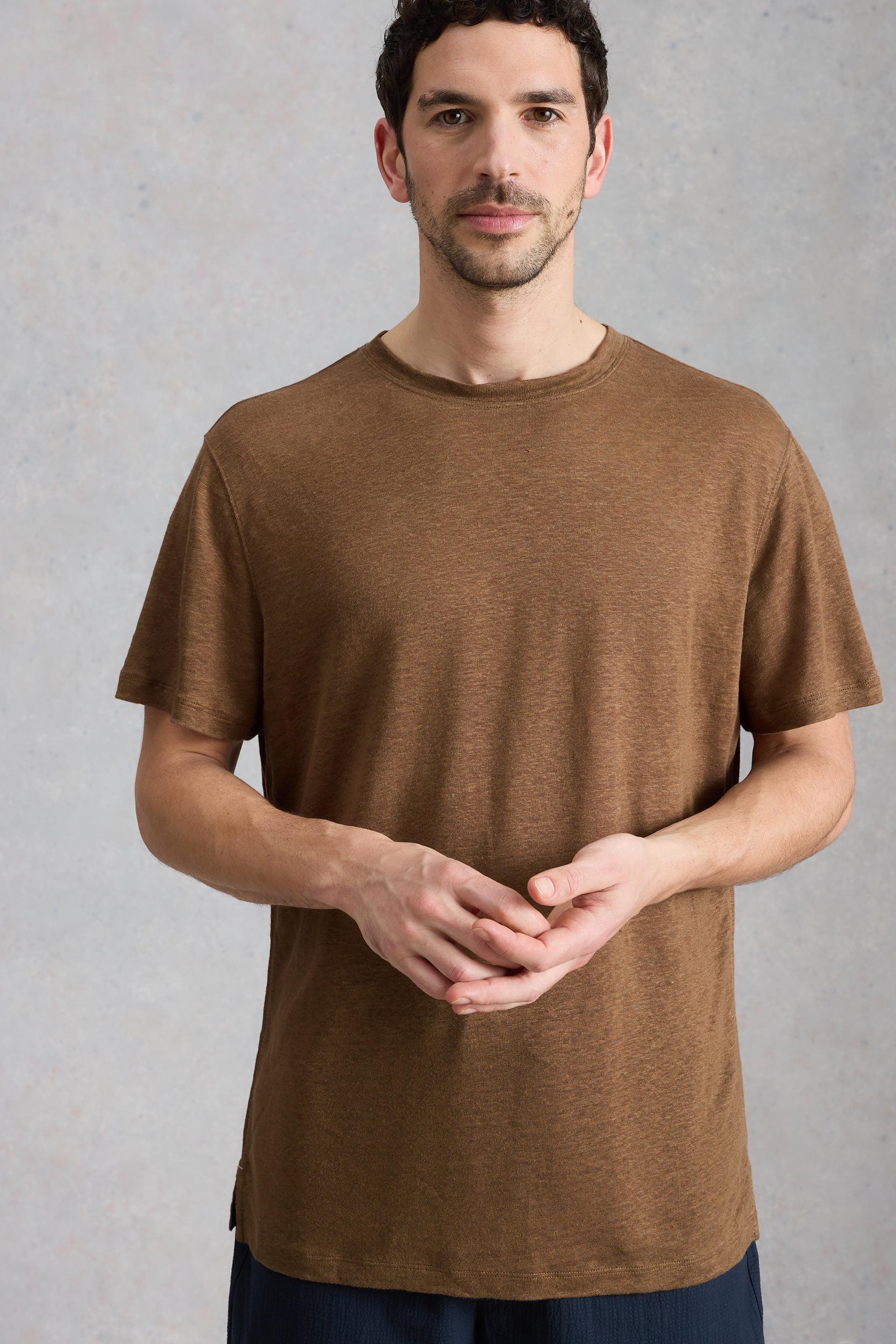A man wearing a brown sustainable mens linen t-shirt, standing against a neutral grey studio background.