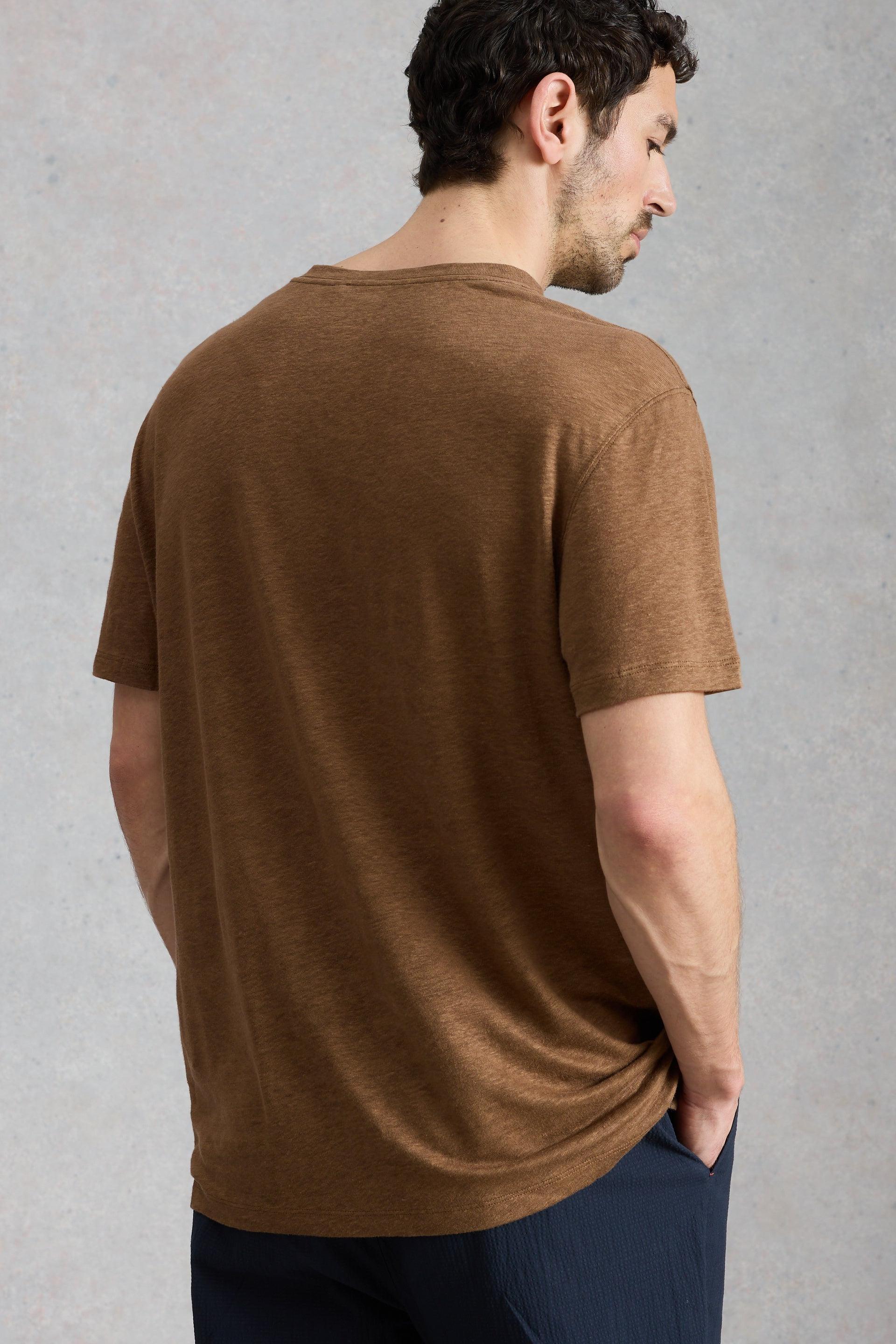 A man wearing a brown mens linen t-shirt, standing against a neutral textured studio background.