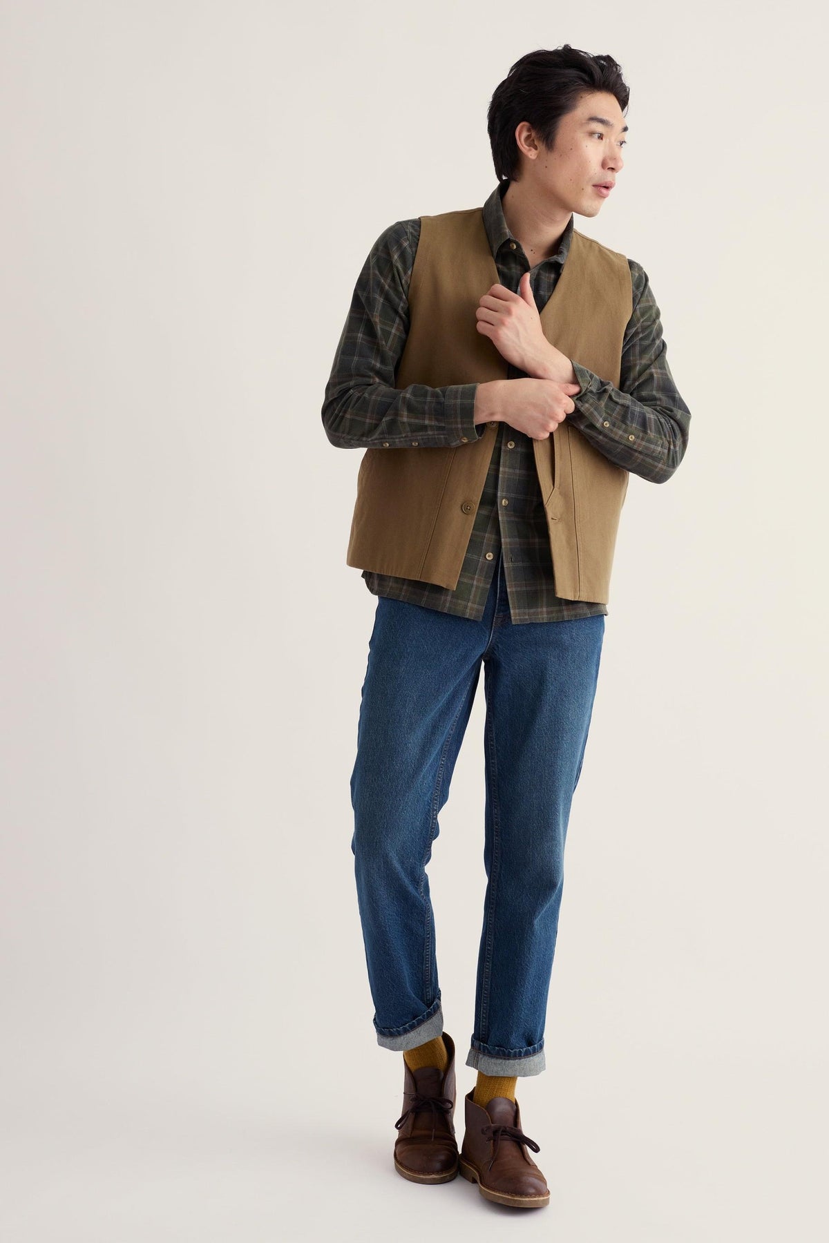 A man modeling a brown suede vest over a dark green plaid shirt, paired with blue jeans, brown shoes, and yellow socks, against a plain light beige studio background.