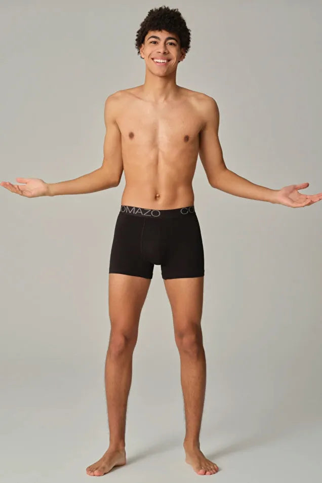 A smiling man wearing black Comazo organic cotton boxer briefs standing against a grey background.