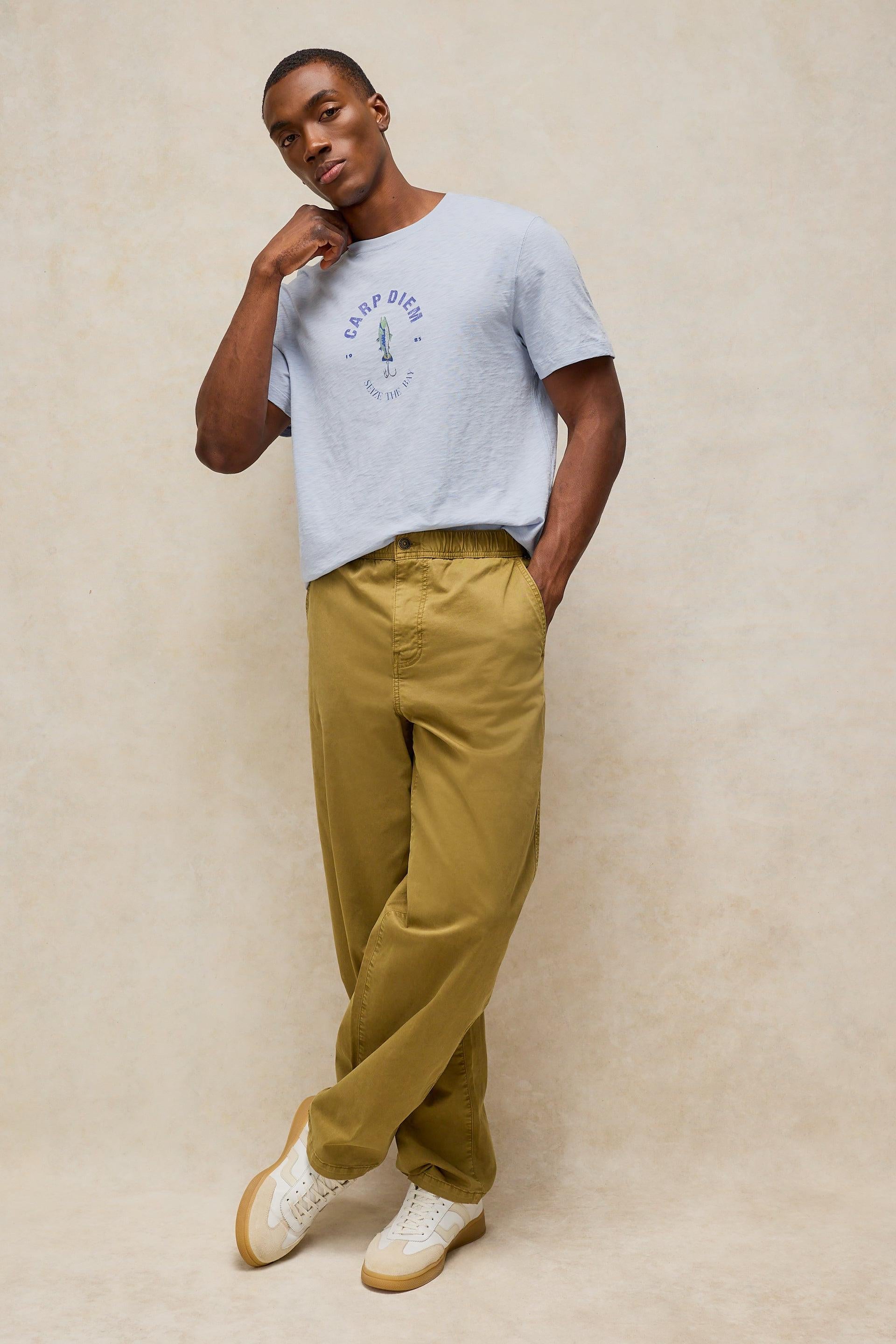 A man modeling a light blue crewneck t-shirt with a 'Carp diem' graphic, paired with olive green trousers and white sneakers, standing against a textured beige studio backdrop.
