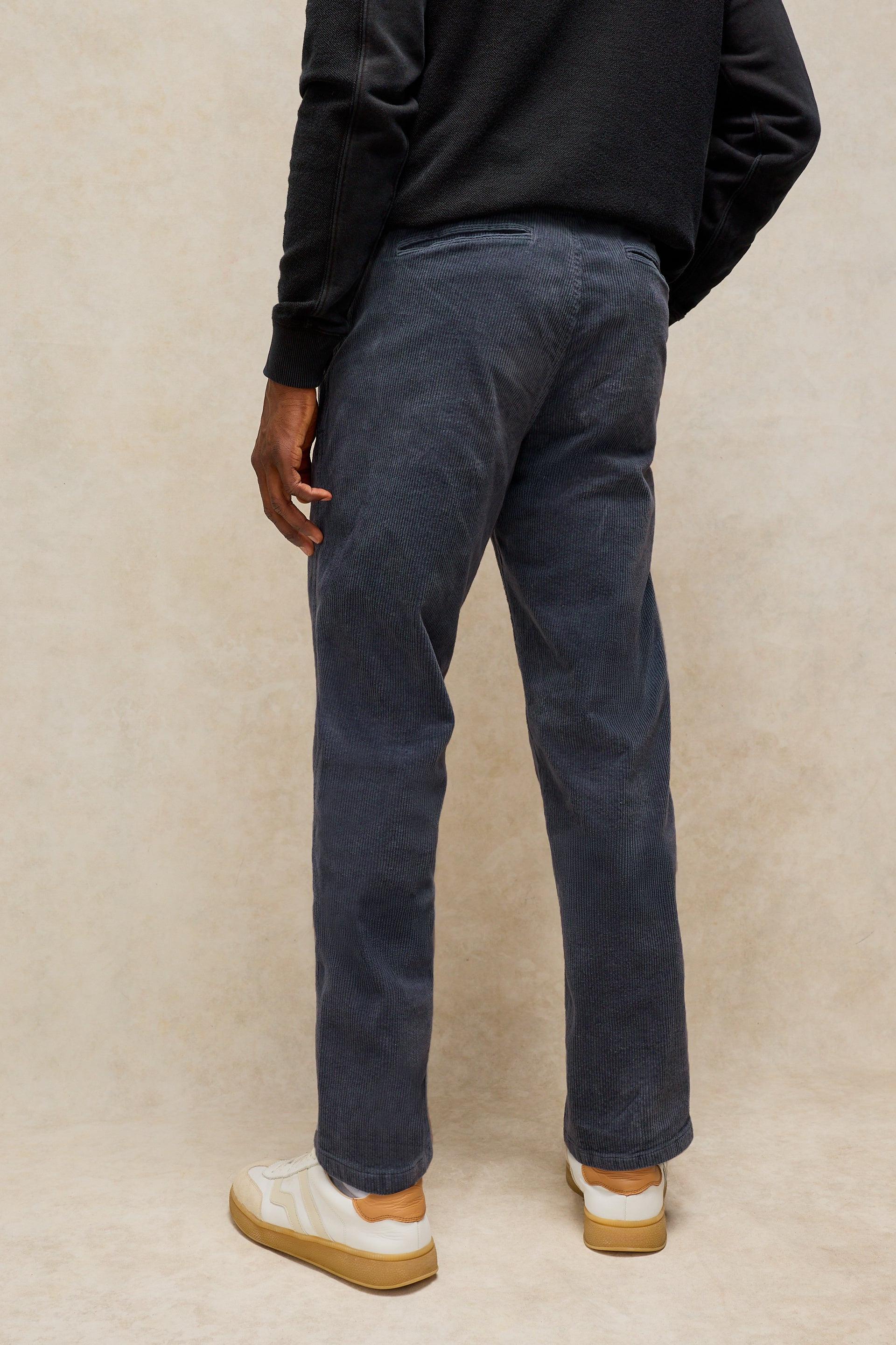 A person shown from the back wearing dark grey corduroy trousers, a black long-sleeve top, and white sneakers with tan soles, standing against a textured beige studio backdrop.