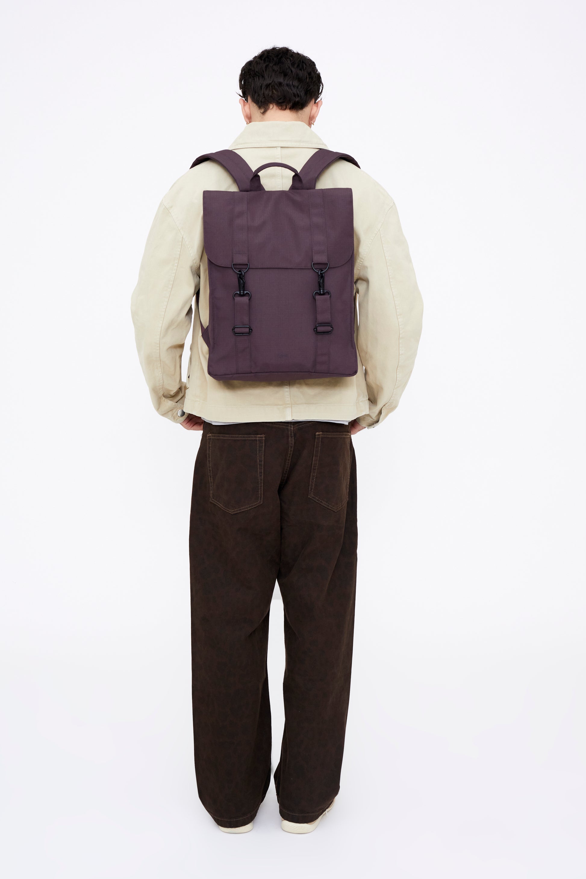 A person viewed from behind wearing a dark purple backpack with dual straps and front buckle details, over a cream jacket and dark brown corduroy pants, against a plain white studio background.