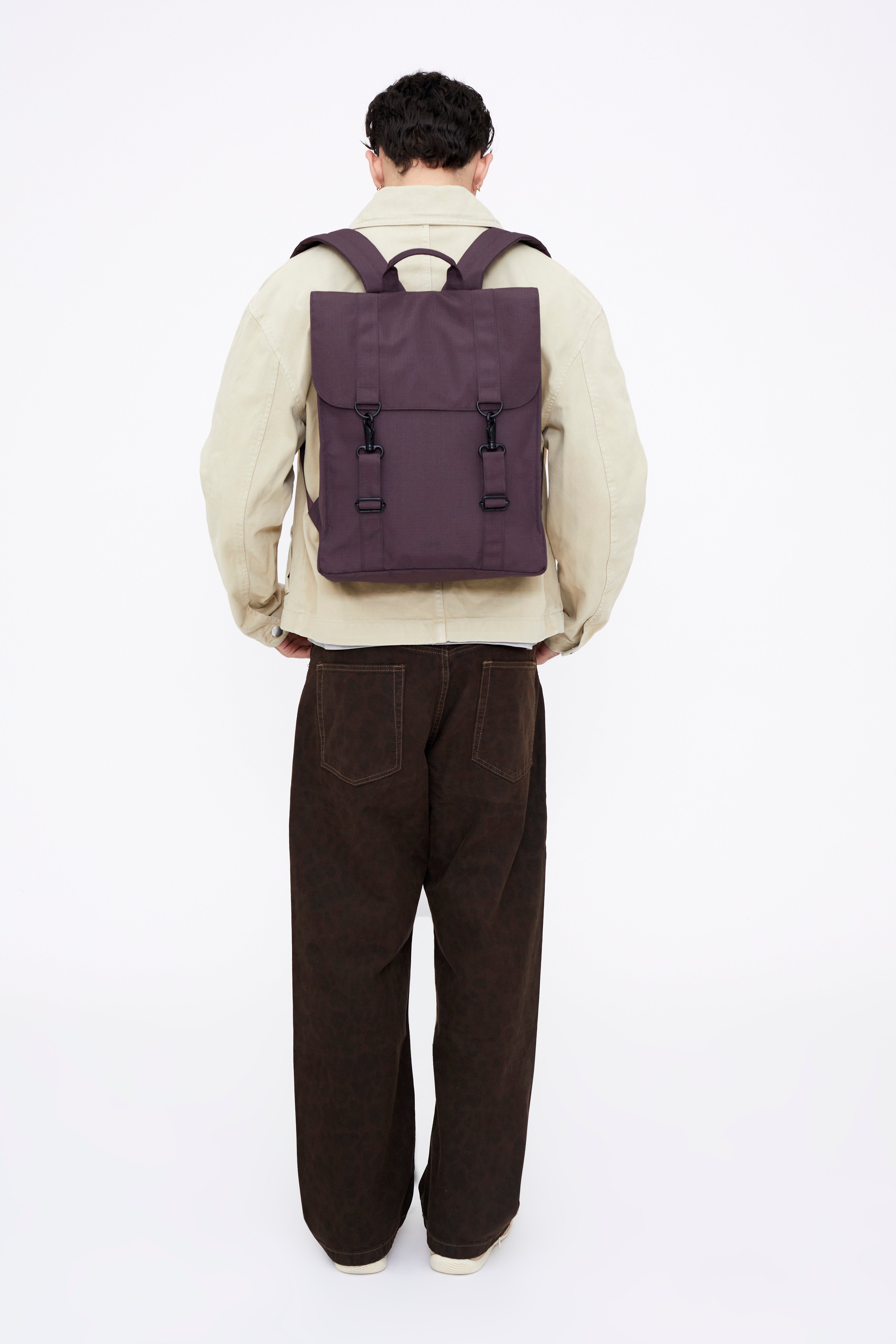 A person viewed from behind wearing a dark purple backpack with dual straps and front buckle details, over a cream jacket and dark brown corduroy pants, against a plain white studio background.