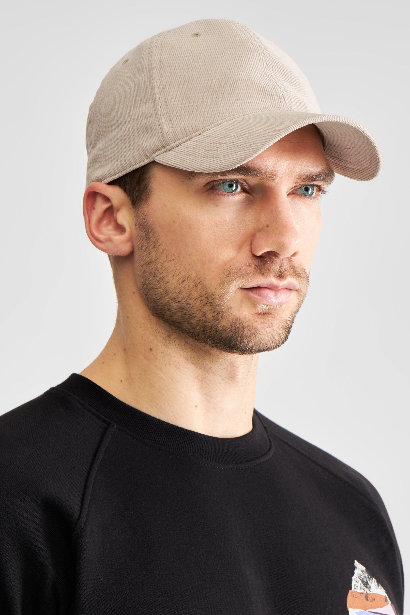 A man wearing a light beige baseball cap and a black raglan sweatshirt, shown in a side profile against a light gray studio background.
