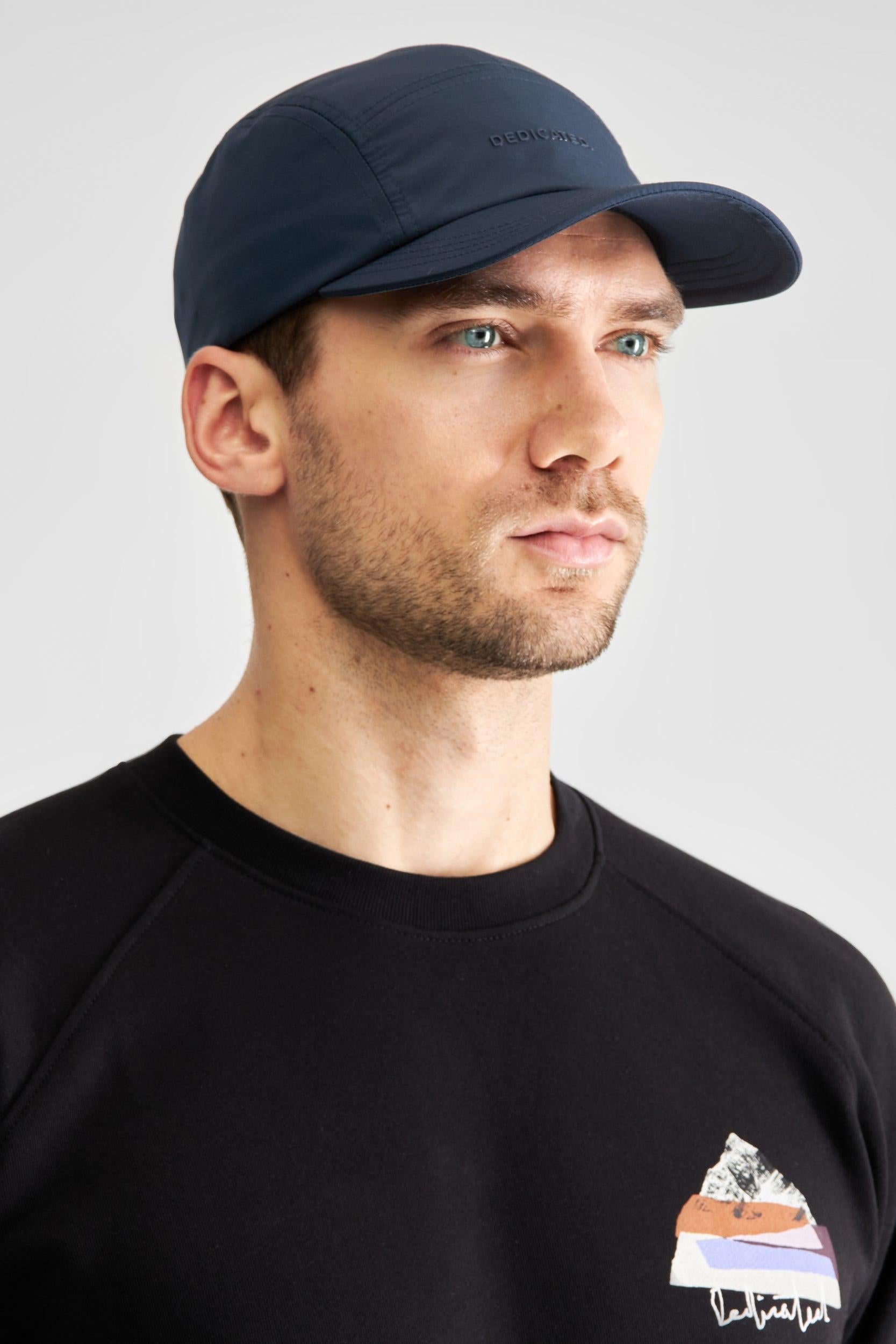 A man wearing a dark navy blue baseball cap and a black Patagonia sweatshirt with a mountain graphic on the chest, photographed against a light gray studio background.