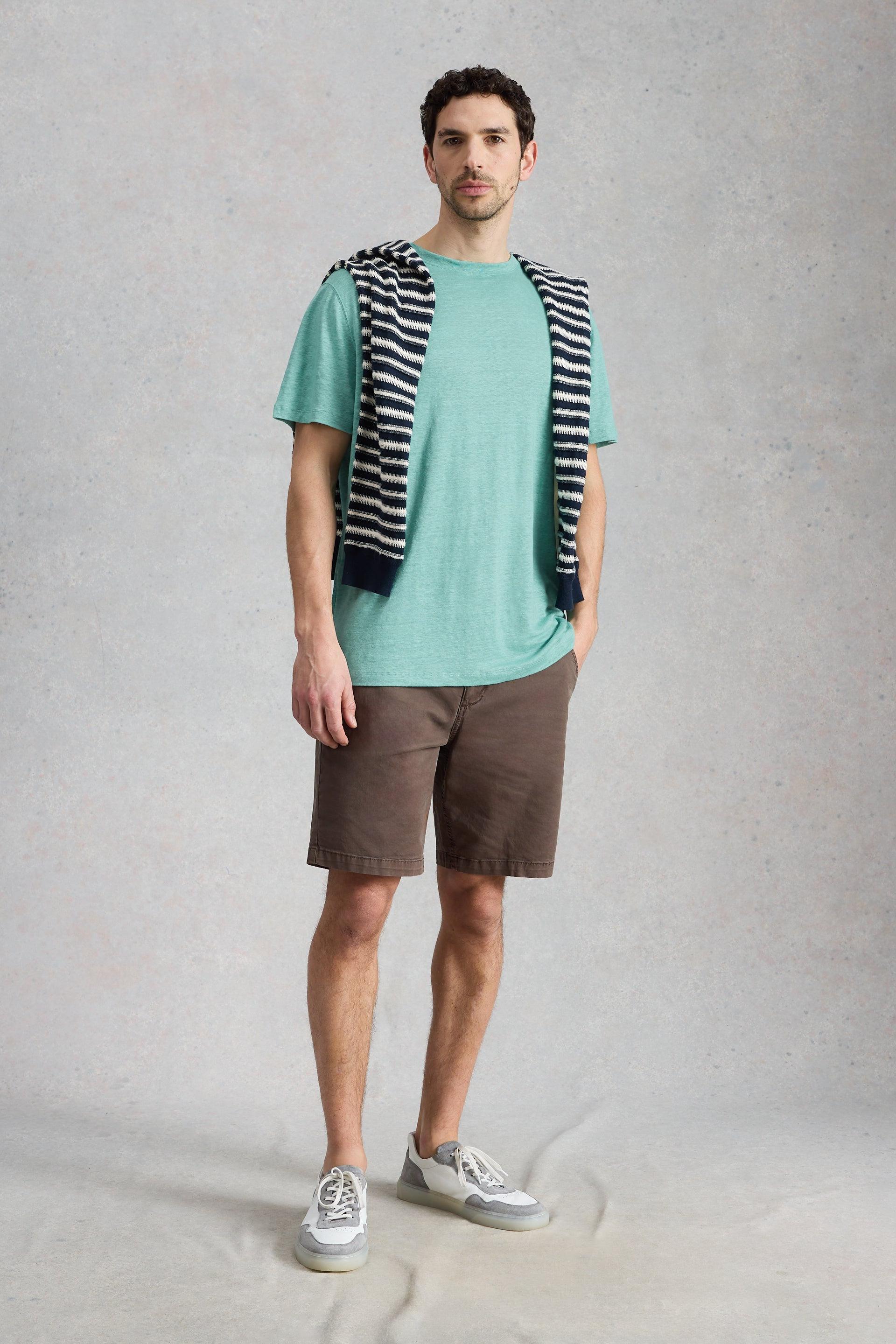 A man wearing a green linen t-shirt with a striped sweater draped over his shoulders and brown shorts.