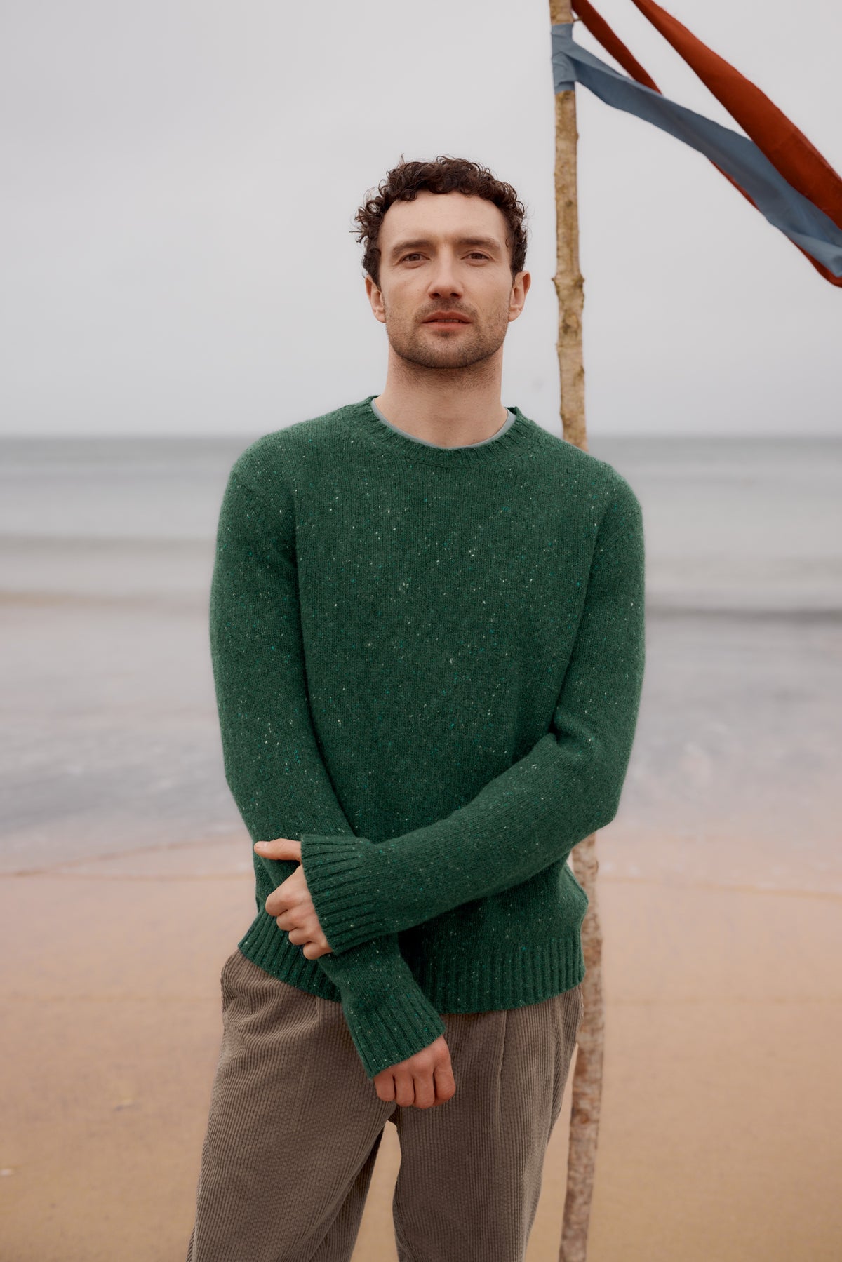 A man wearing a green merino wool sweater standing on a beach with a flag pole in the background.