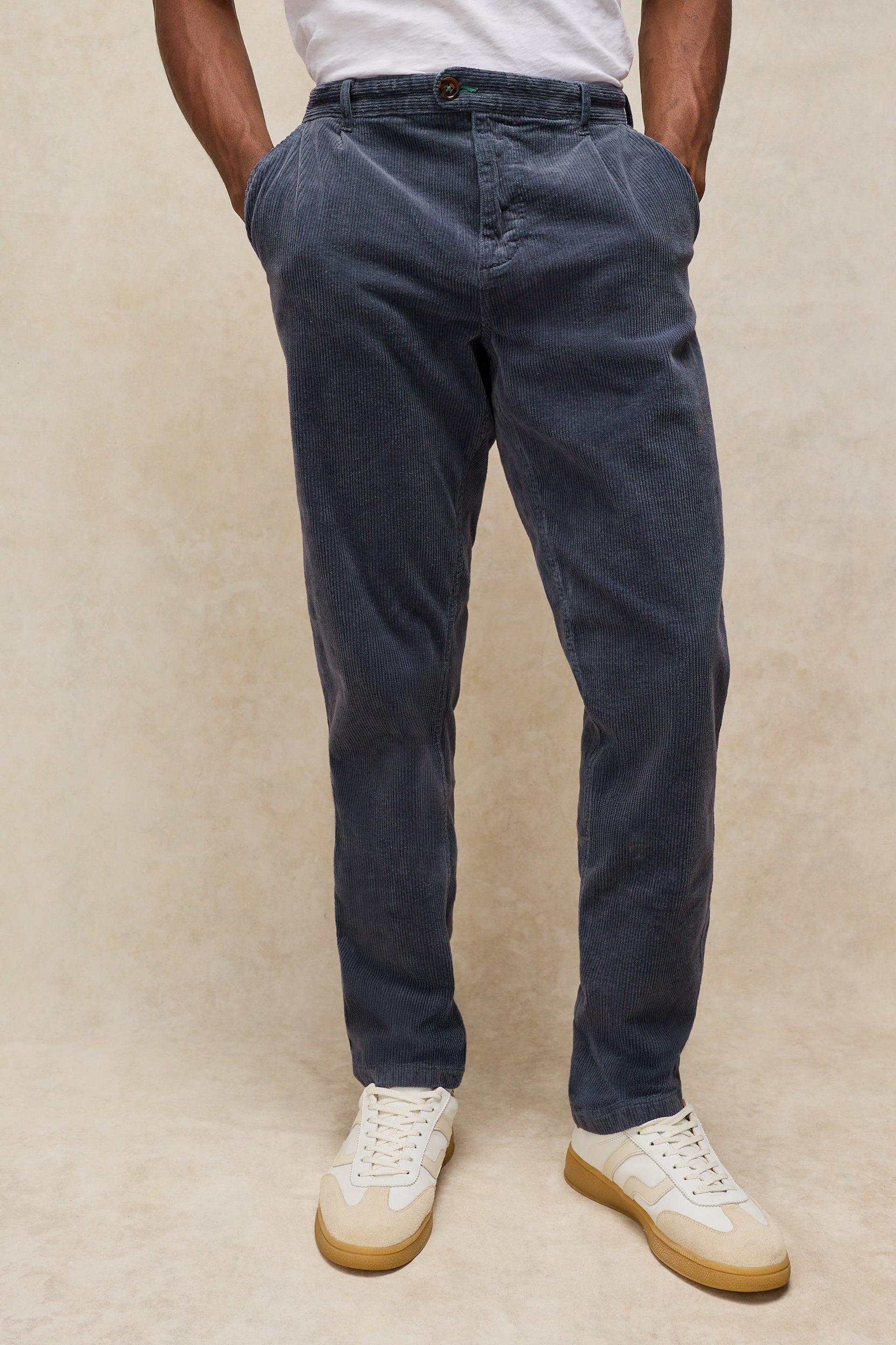 A person wearing slim-fit dark grey corduroy trousers, paired with a white t-shirt and white sneakers with tan soles, standing against a textured beige background.