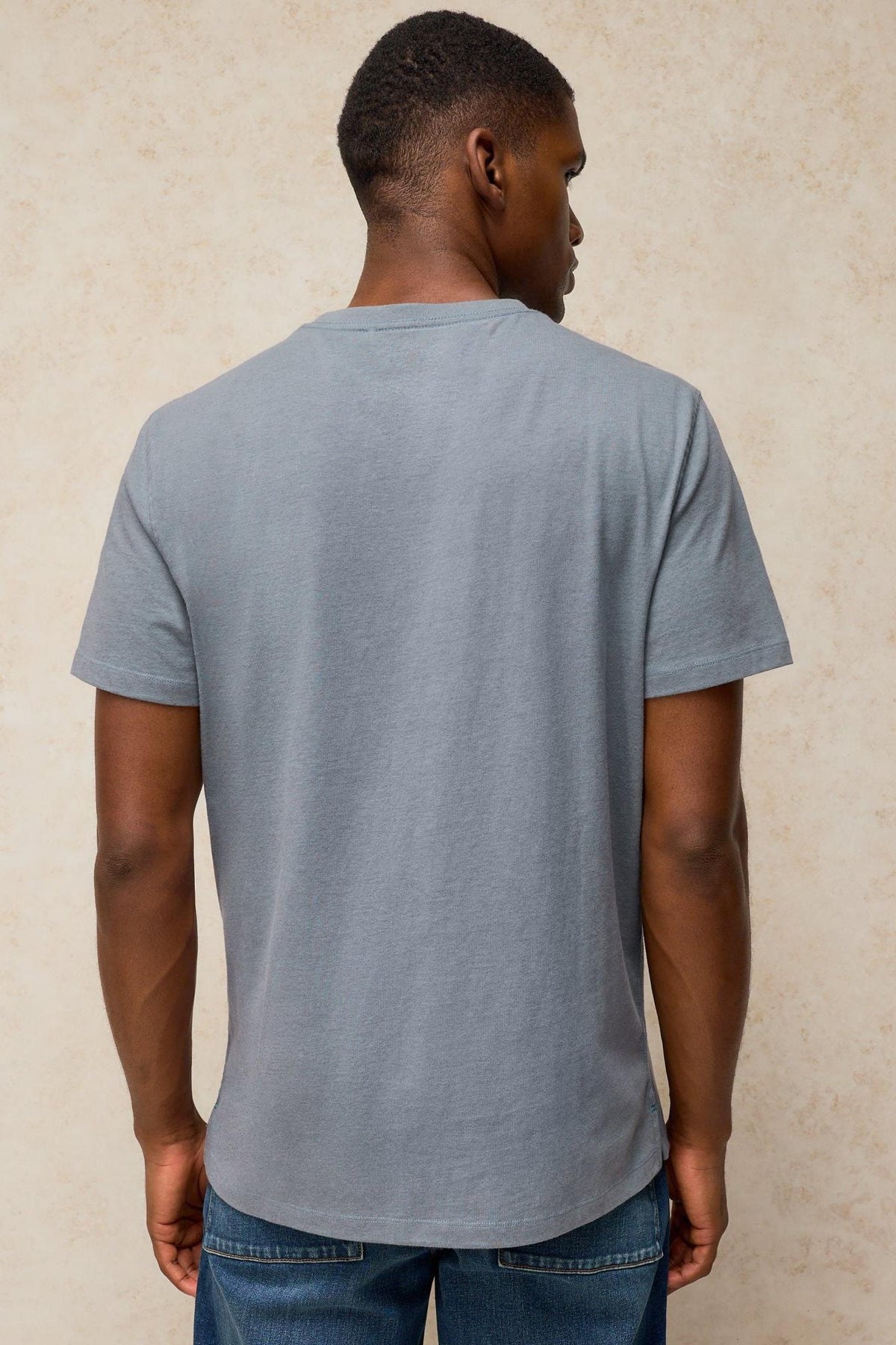 Rear view of a man wearing a plain grey organic cotton t-shirt against a textured beige background.