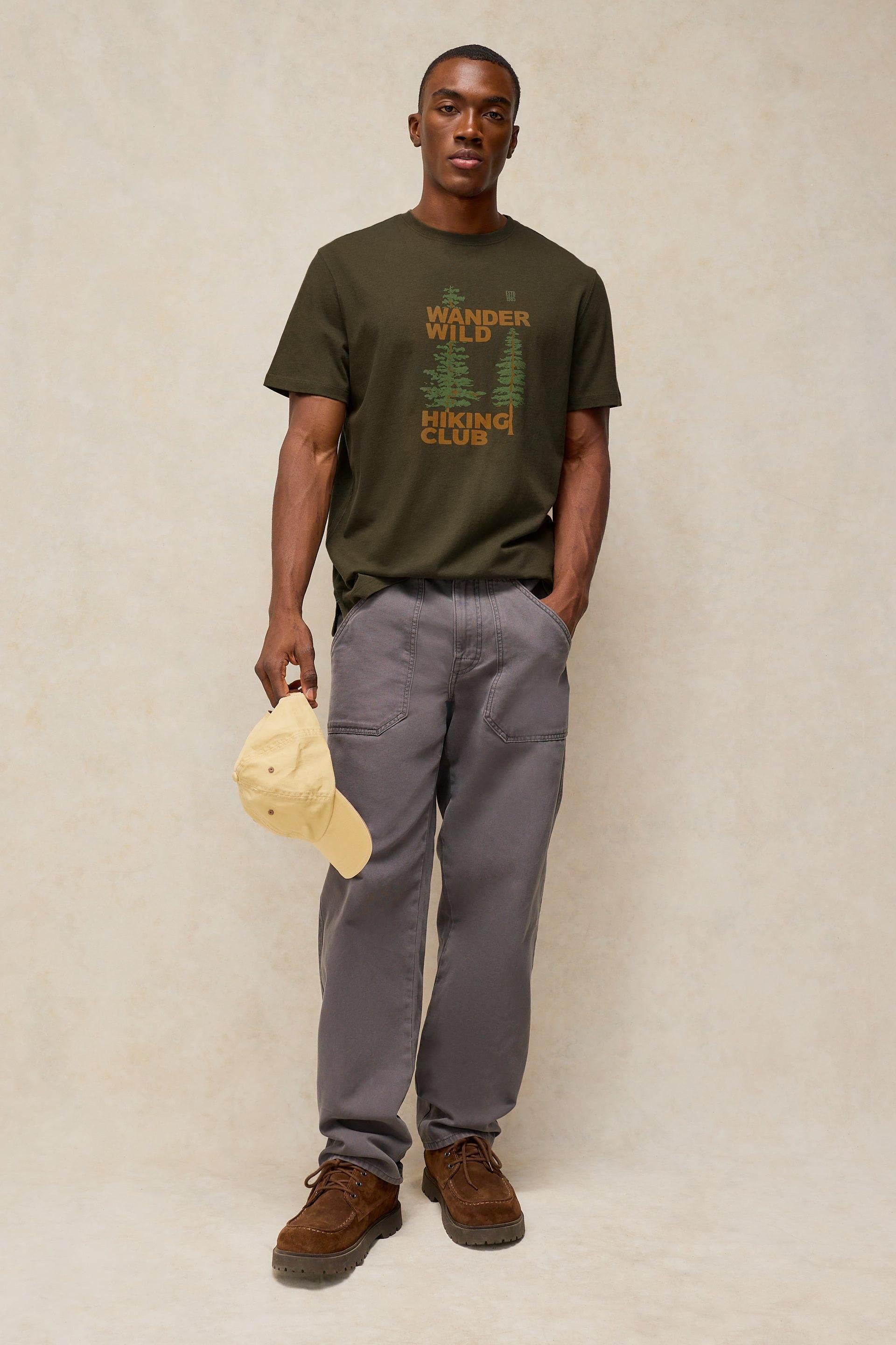 A man wearing an olive Wander Wild Hiking Club t-shirt and grey pants holds a yellow cap against a plain studio backdrop