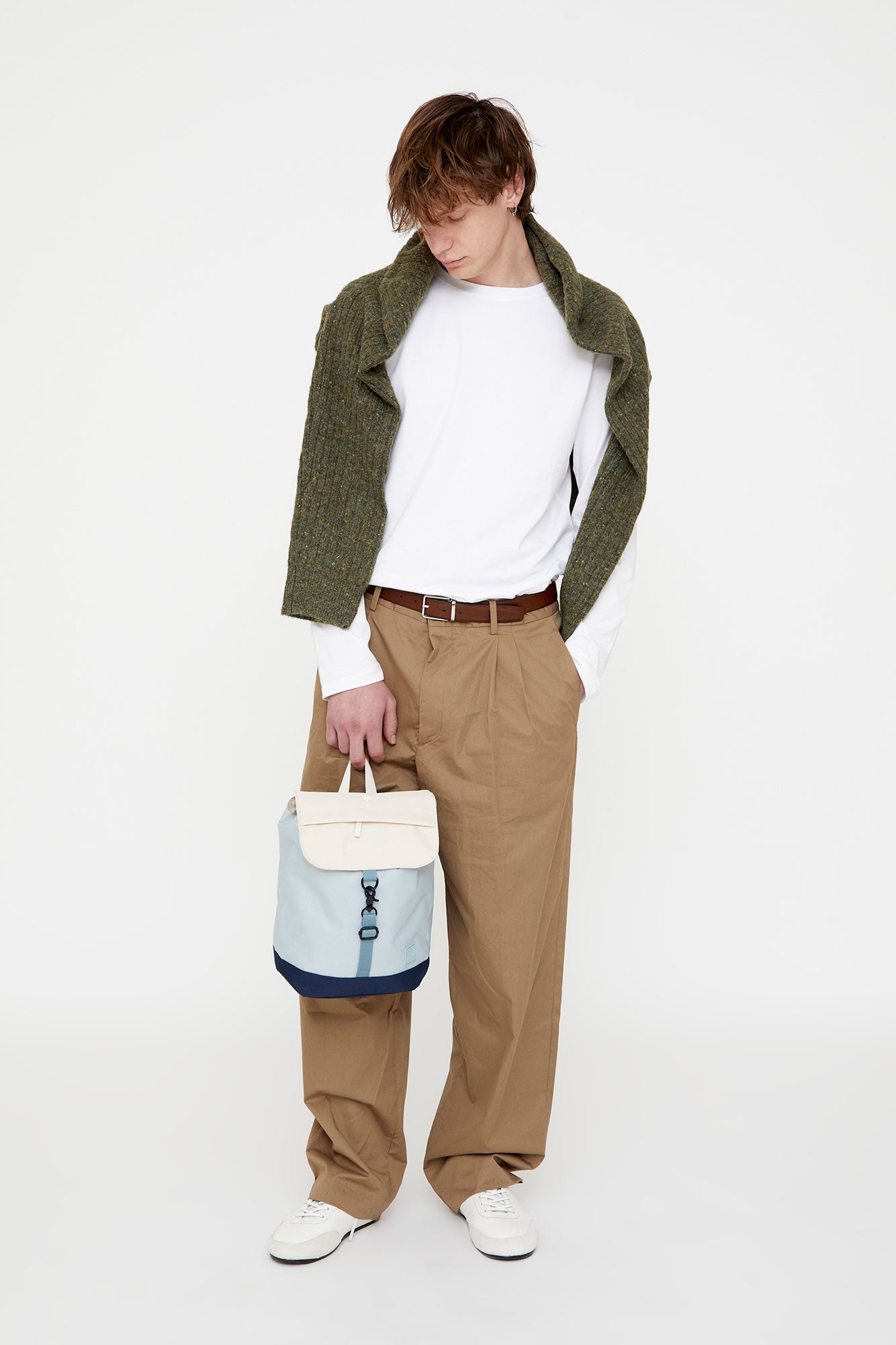 Person wearing a green jacket, white shirt, and beige pants, holding a blue and white bag on a white background