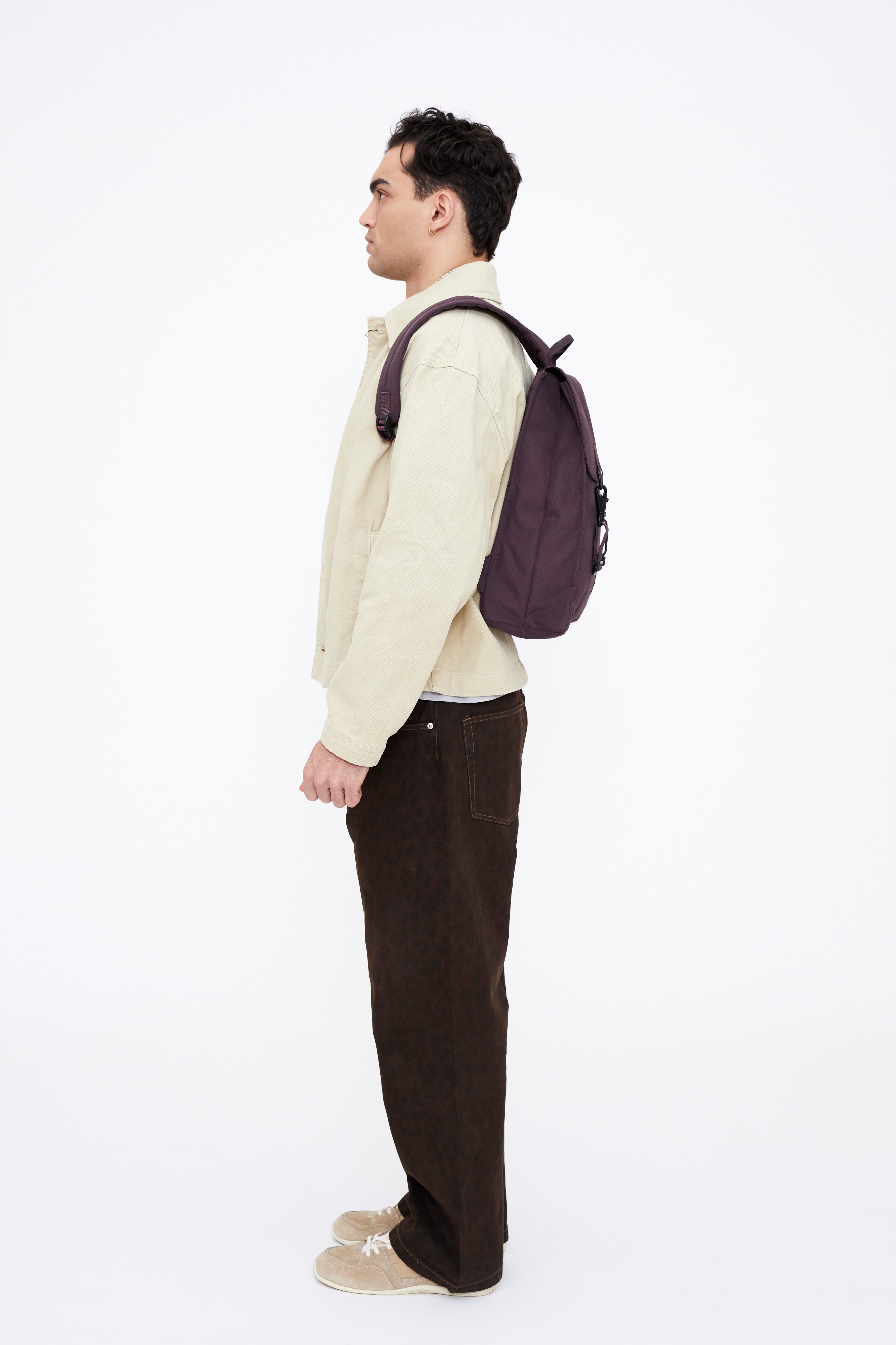 A man in a cream jacket, dark brown corduroy pants, and beige sneakers stands in profile wearing a dark purple backpack against a plain white background.