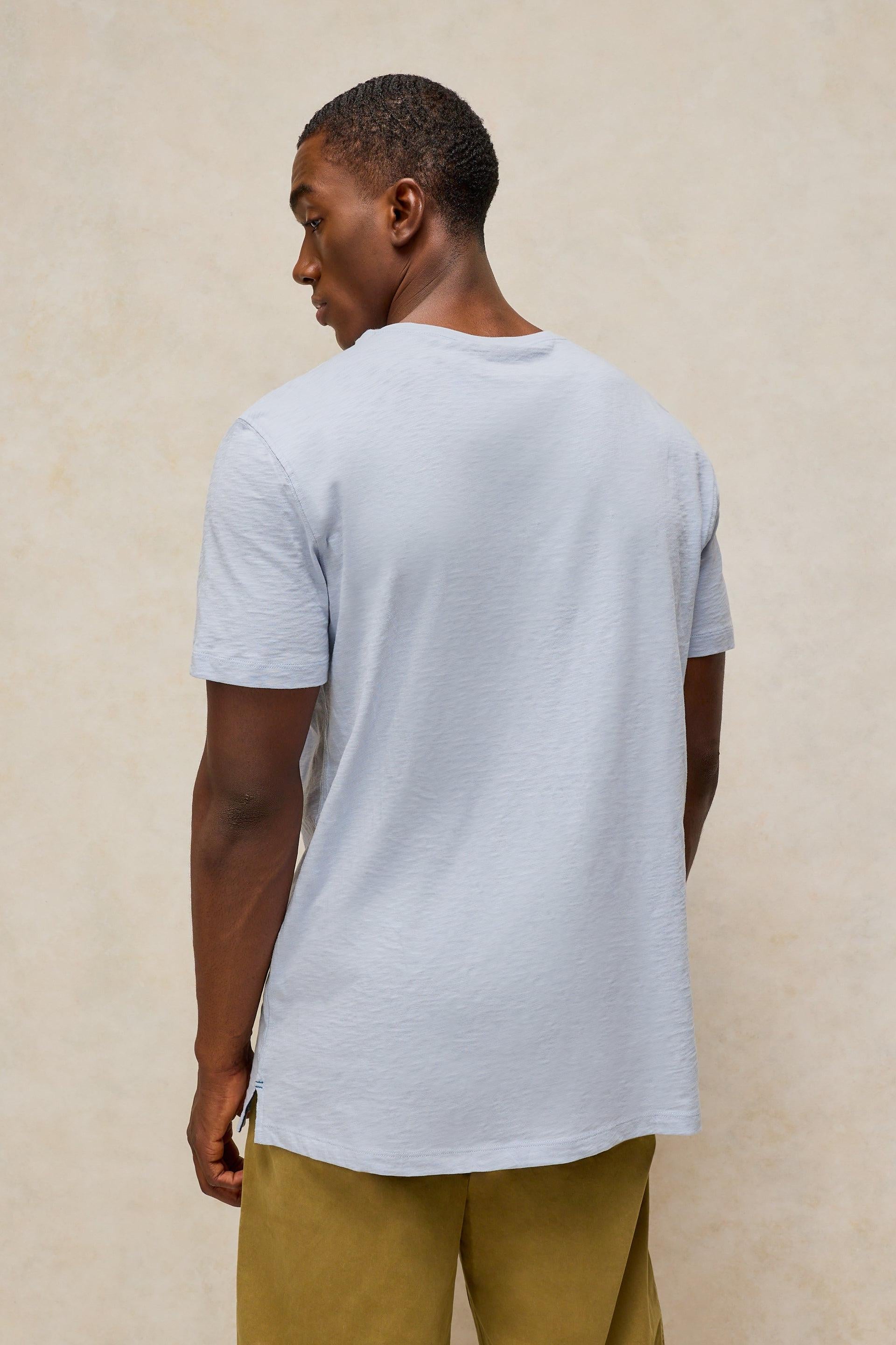 A man wearing a light blue-gray short-sleeve t-shirt and olive green pants, shown from a back-three-quarter view against a textured beige wall.