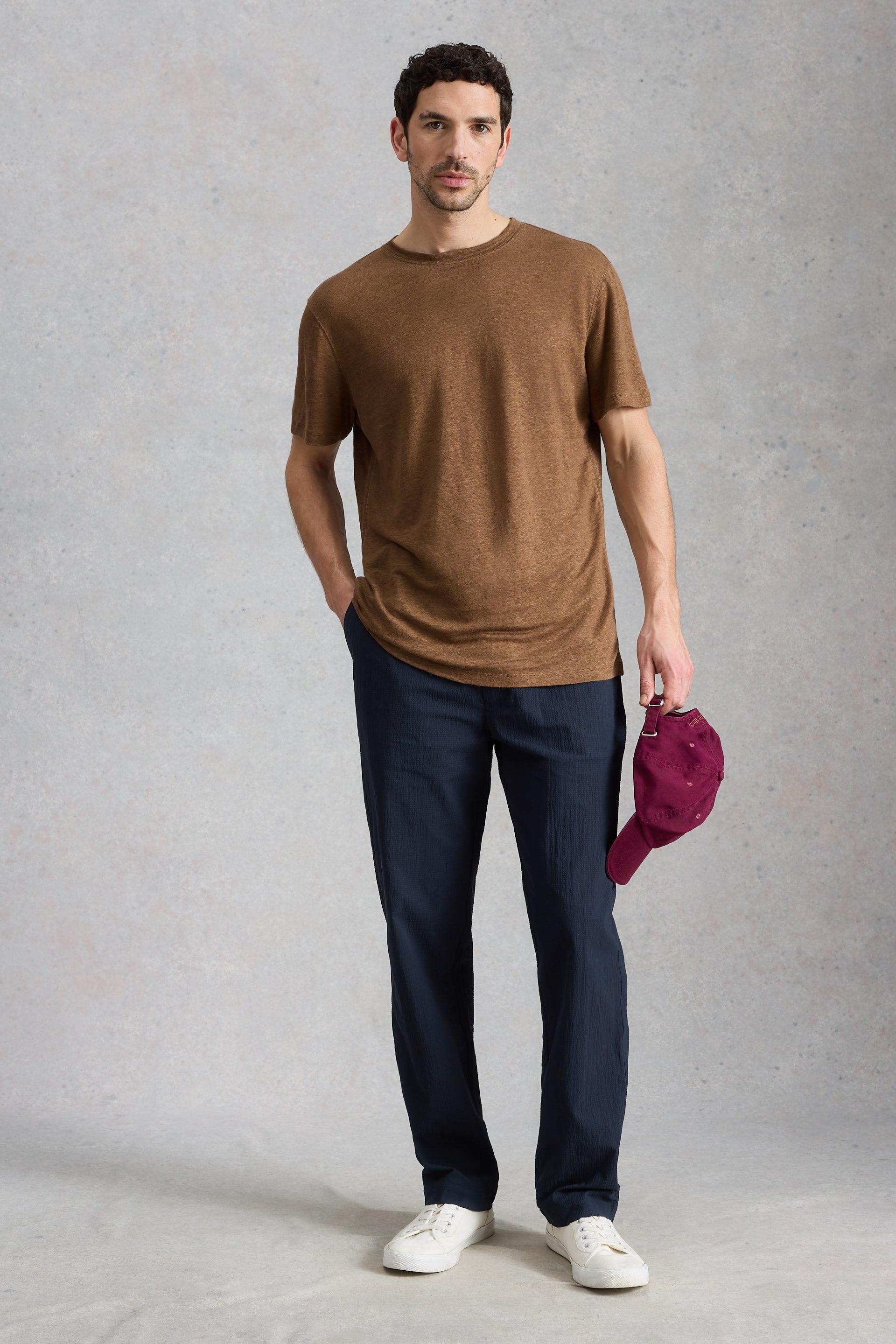 A man wearing a brown mens linen t-shirt and navy trousers, holding a maroon cap against a neutral studio background.