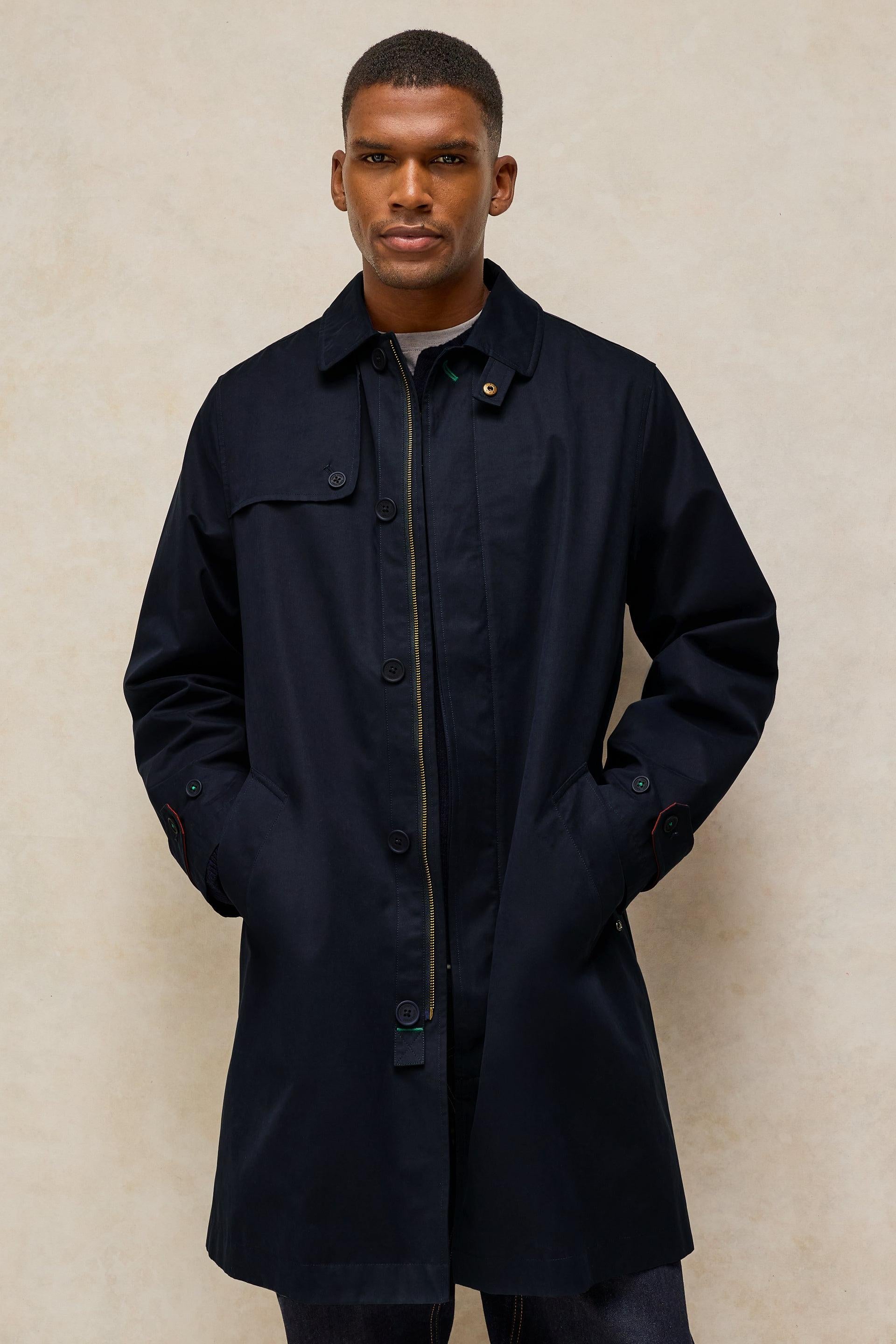 A man wearing a dark navy blue mid-length coat with a front zipper and flap chest pockets, standing against a textured beige wall.