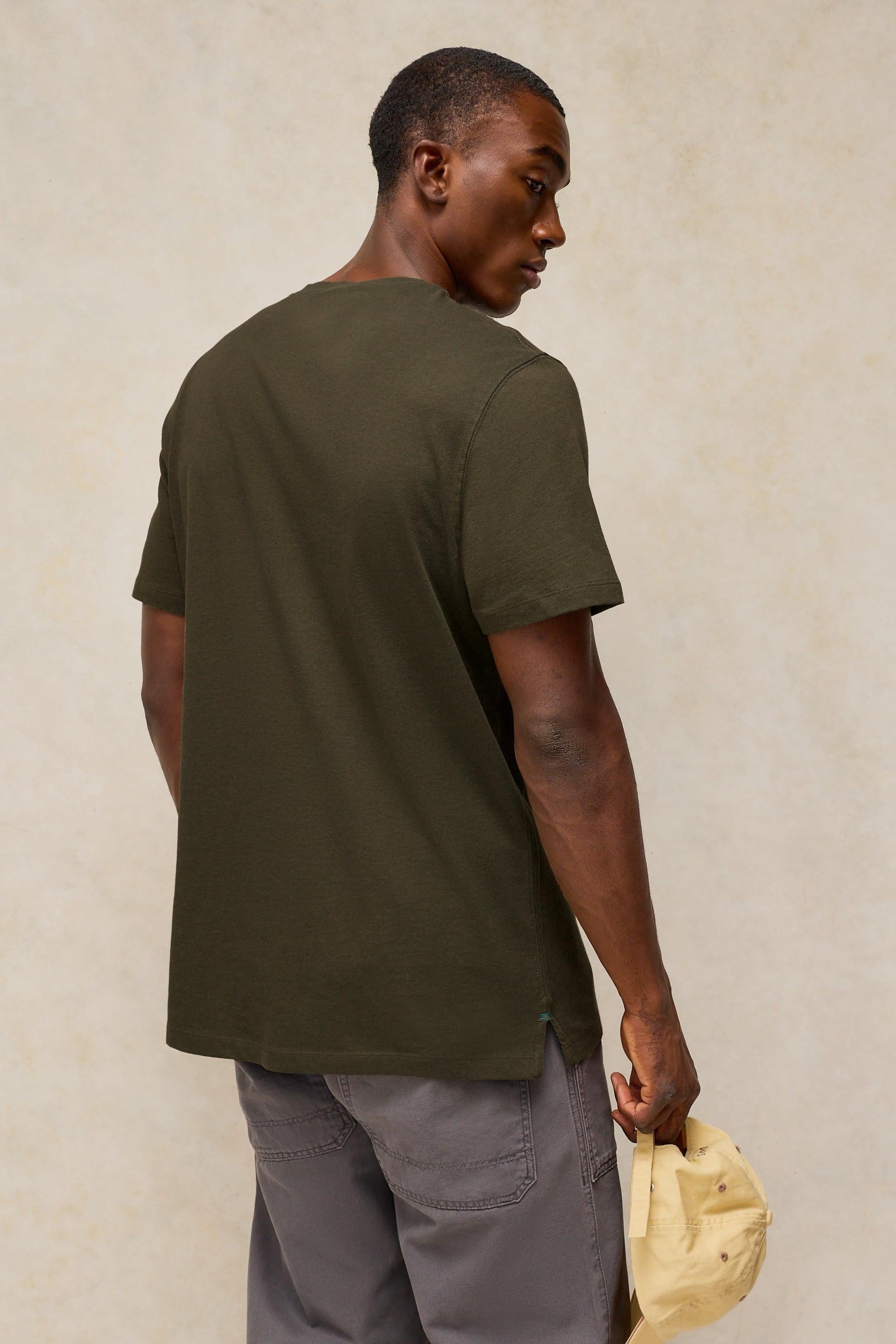 A man viewed from behind wearing an olive green cotton t-shirt and grey pants while holding a beige baseball cap