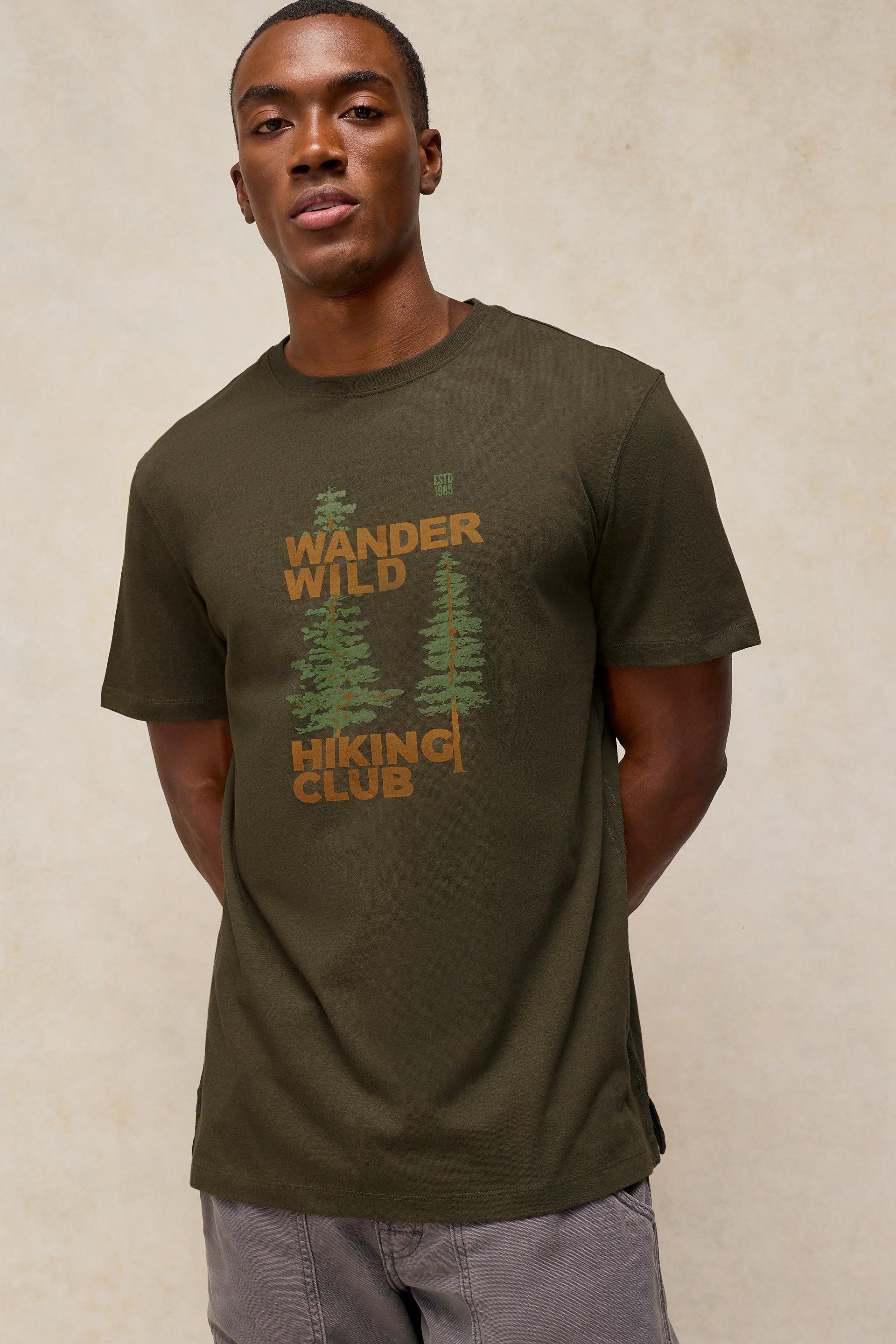 A man wearing an olive green t-shirt with Wander Wild Hiking Club graphic design and forest illustration
