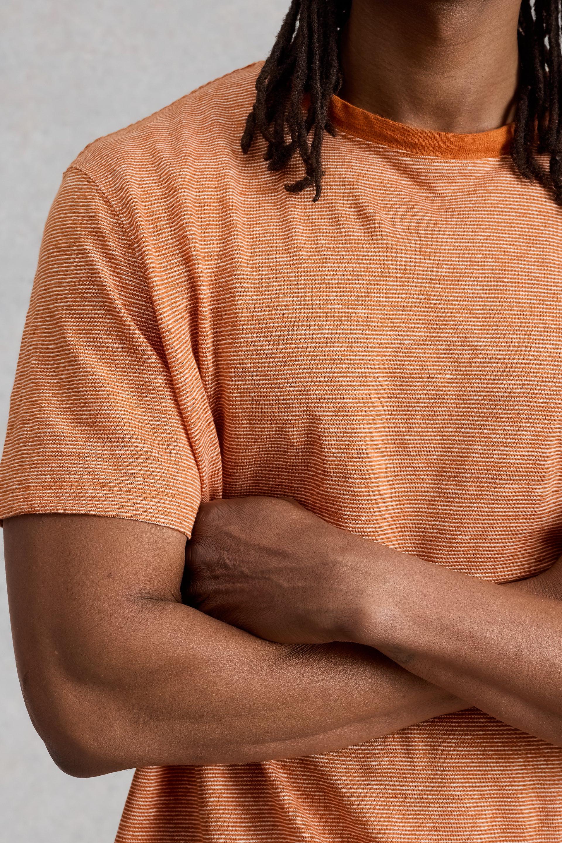Close-up of a man wearing an orange and white striped 100% linen t-shirt with arms crossed.