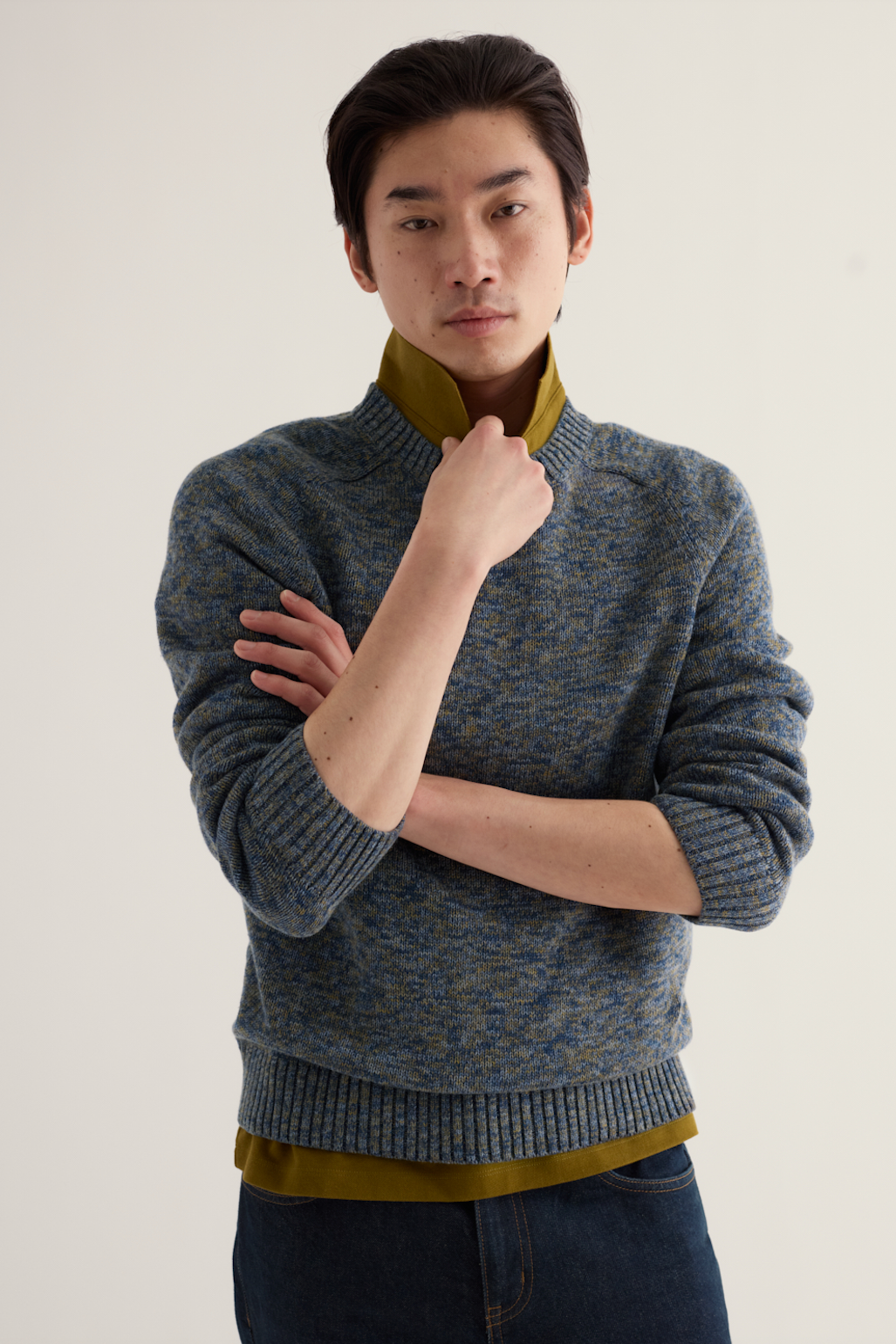 A man wearing a blue and gray marled knit sweater over a mustard yellow collared shirt, paired with dark blue jeans, standing against a plain off-white background.