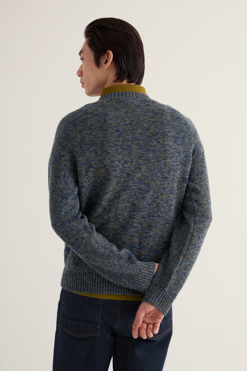 A man shown from the back wearing a textured blue and olive green knit sweater over a matching olive green shirt, paired with dark blue jeans, against a plain off-white background.