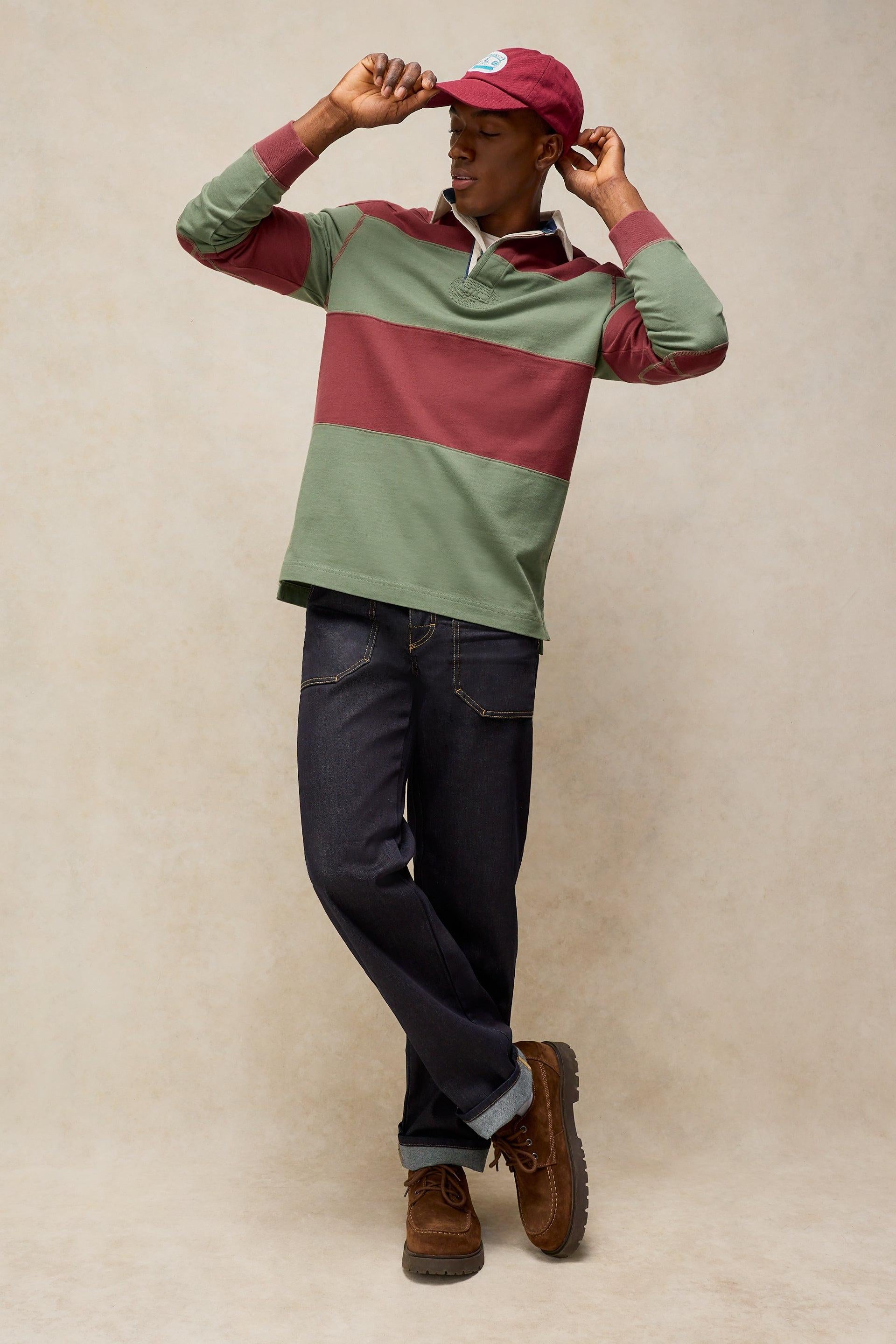 A man modeling a color-blocked long-sleeve rugby shirt with green and dusty red horizontal stripes, paired with dark blue jeans and brown boots, against a neutral beige studio background.