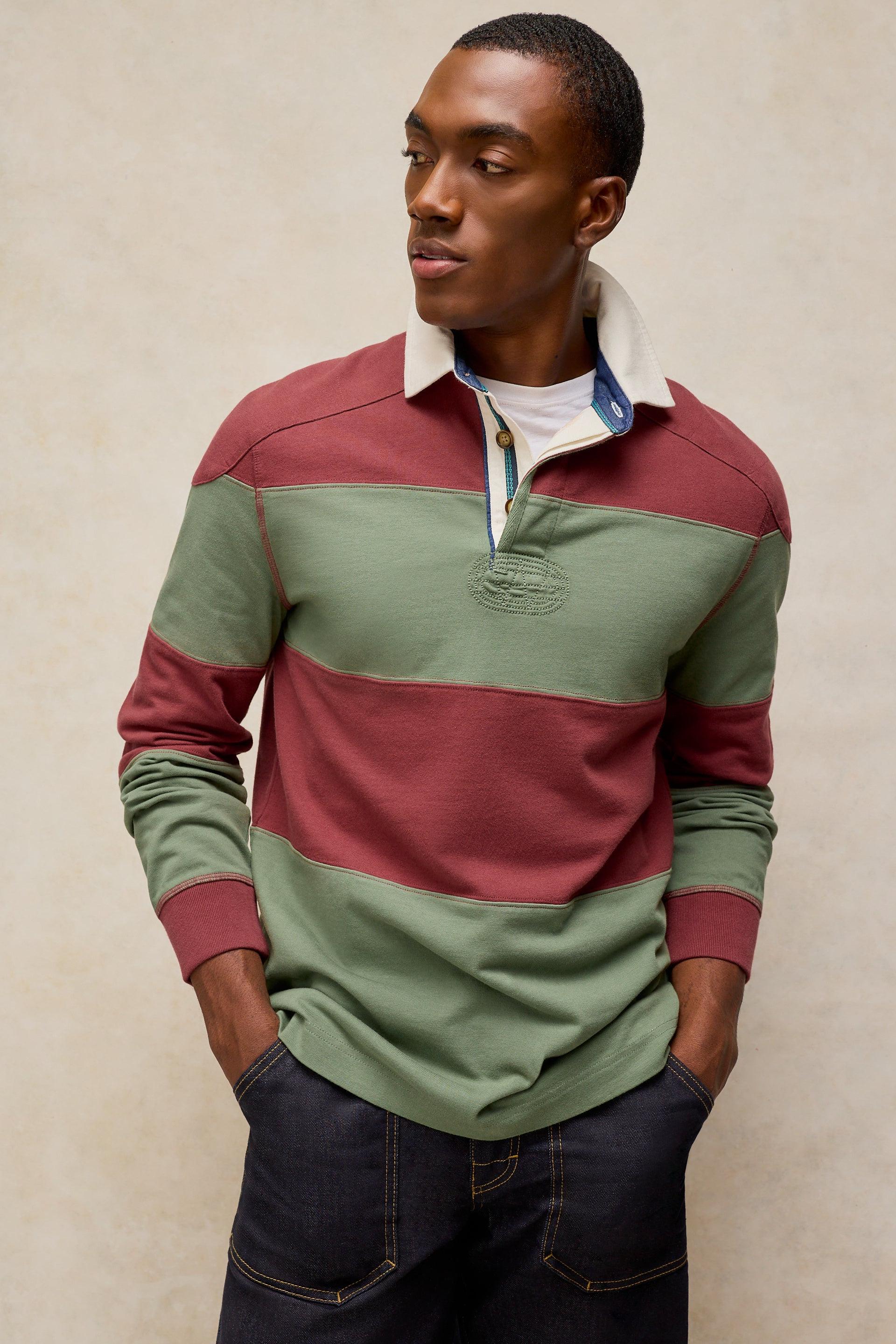 A man wearing a color-blocked long-sleeve rugby shirt with horizontal stripes of sage green, burgundy, and white, paired with dark jeans, standing against a textured beige wall.