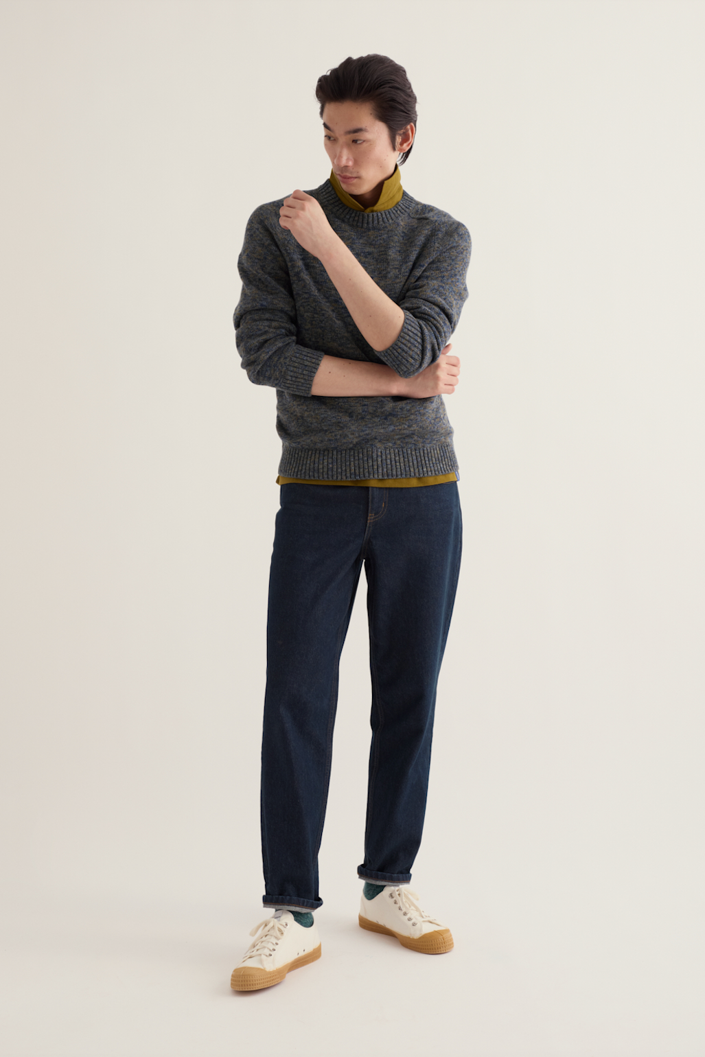 A male model stands against a plain light beige studio background wearing a dark gray knit sweater over a mustard yellow turtleneck, dark blue jeans, and cream sneakers with tan soles.