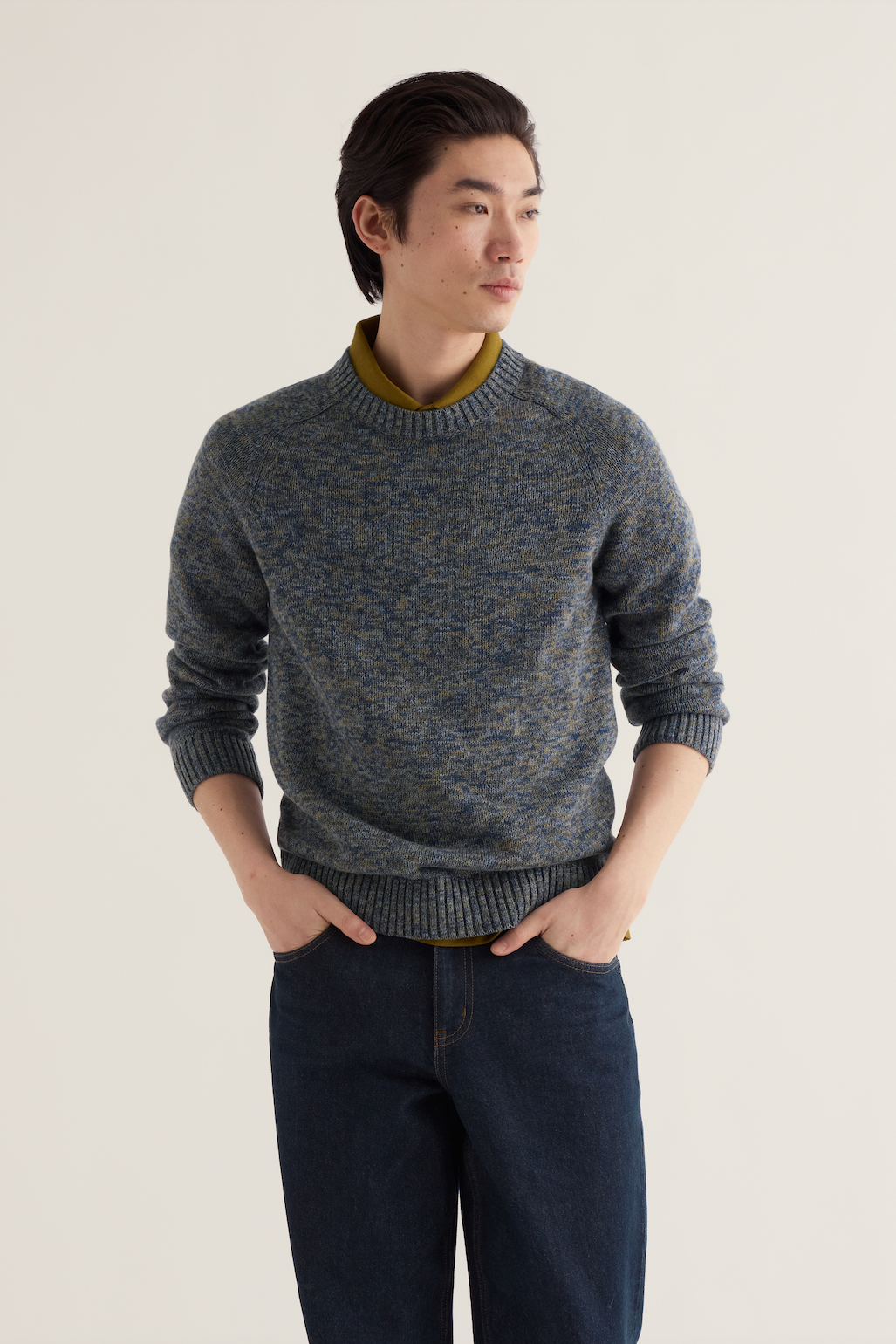 A man wearing a marled blue and gray knit sweater with a contrasting olive green collar, paired with dark blue jeans, standing against a plain light beige background.
