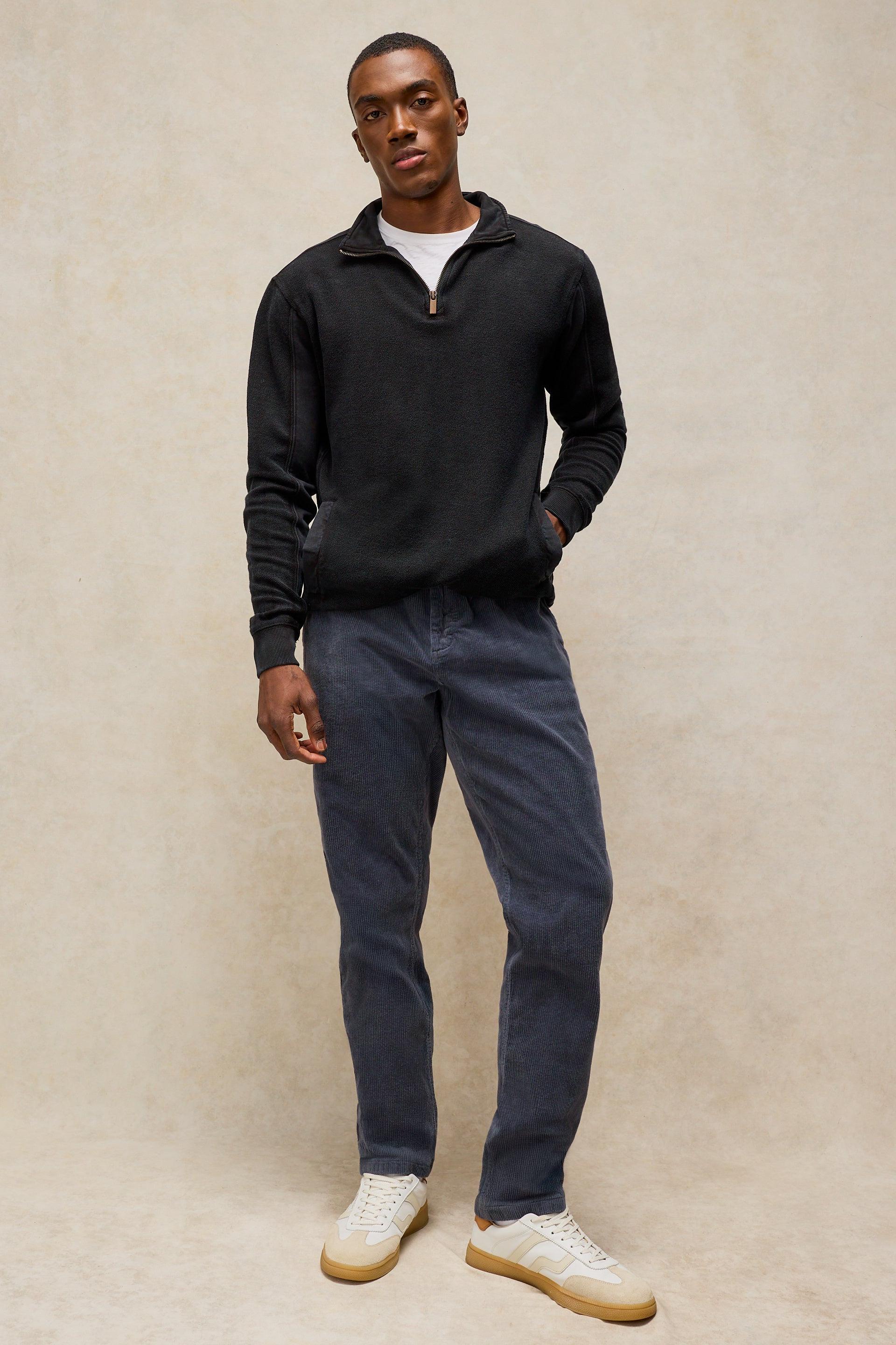 A man modeling a black long-sleeve quarter-zip pullover sweater, paired with dark grey trousers and white sneakers, standing against a textured beige studio backdrop.
