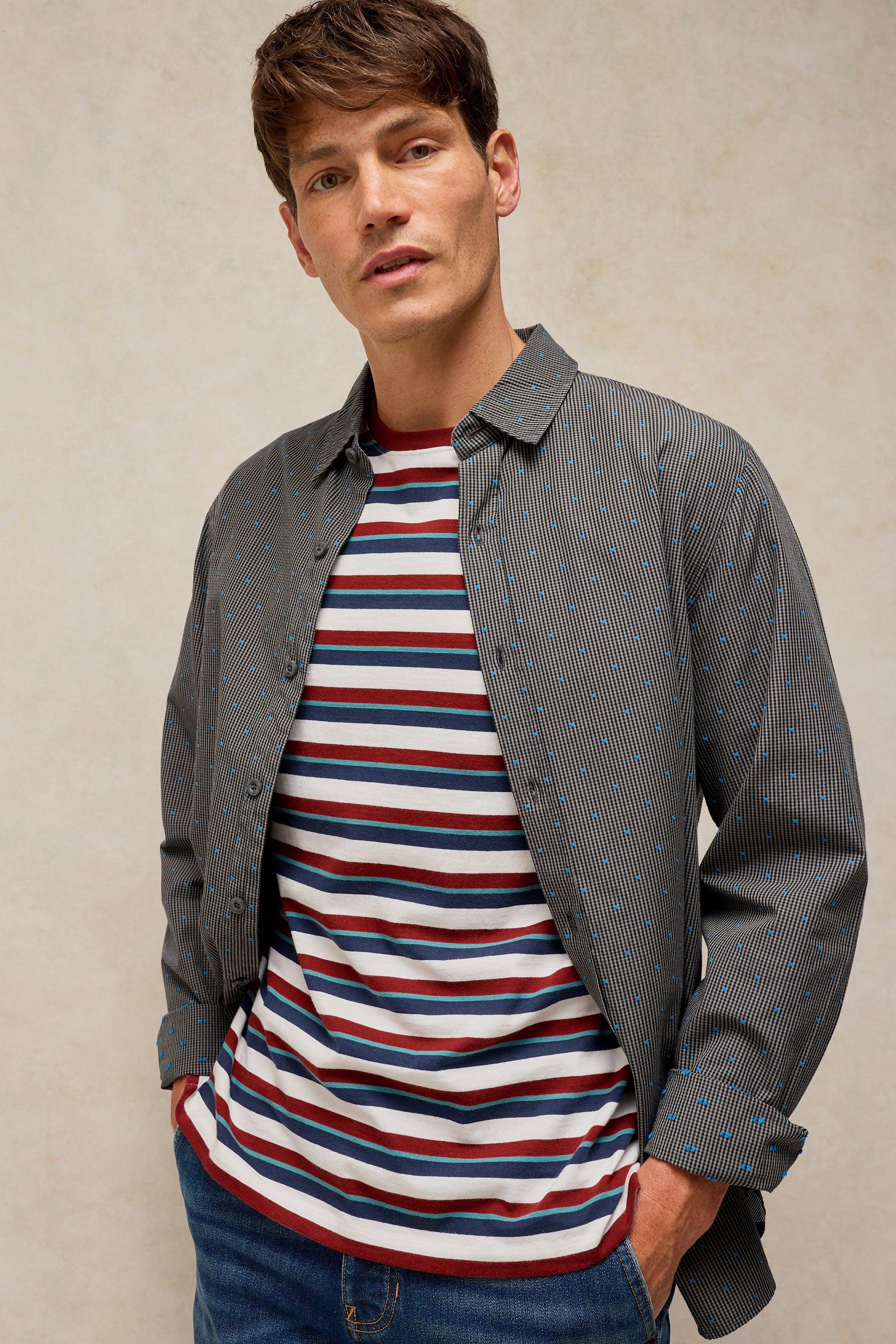 A man wearing a dark gray pinstriped button-up shirt over a red, white, and navy blue striped t-shirt, paired with blue jeans, against a textured beige background.