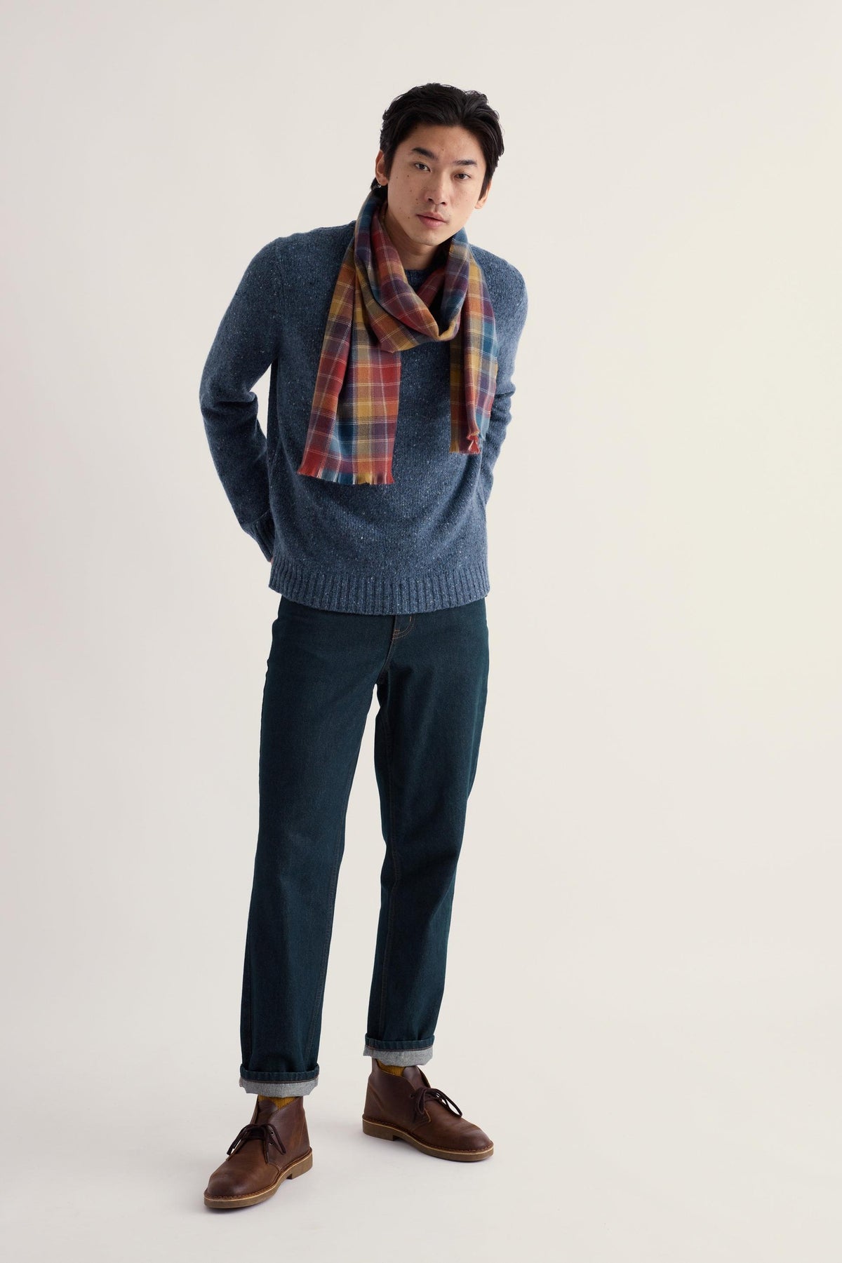 A young man wearing a blue Seasalt merino wool jumper and a plaid scarf against a plain studio background.