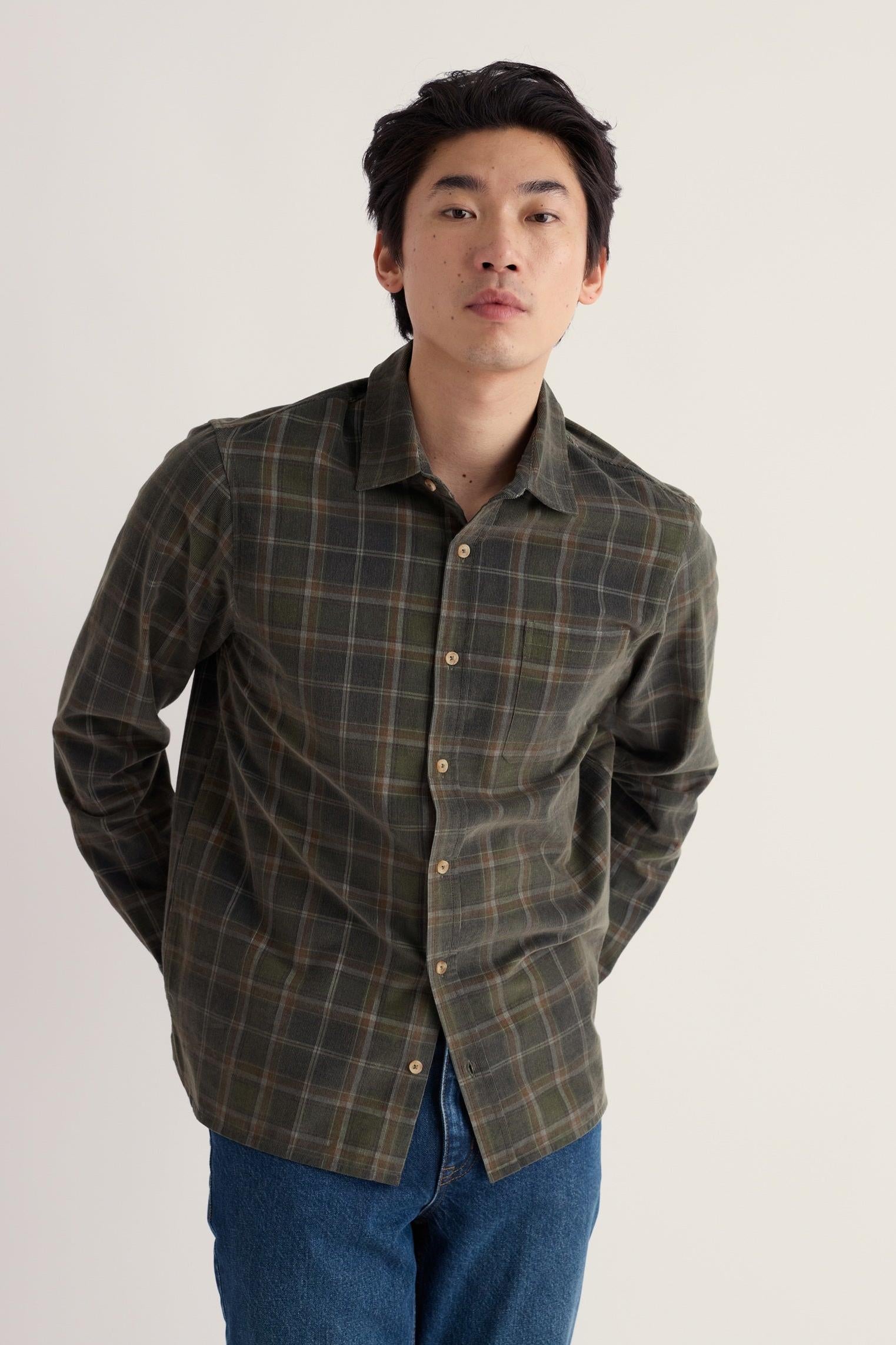 A man wearing a dark olive green plaid button-down shirt with blue jeans, standing against a plain off-white studio background.