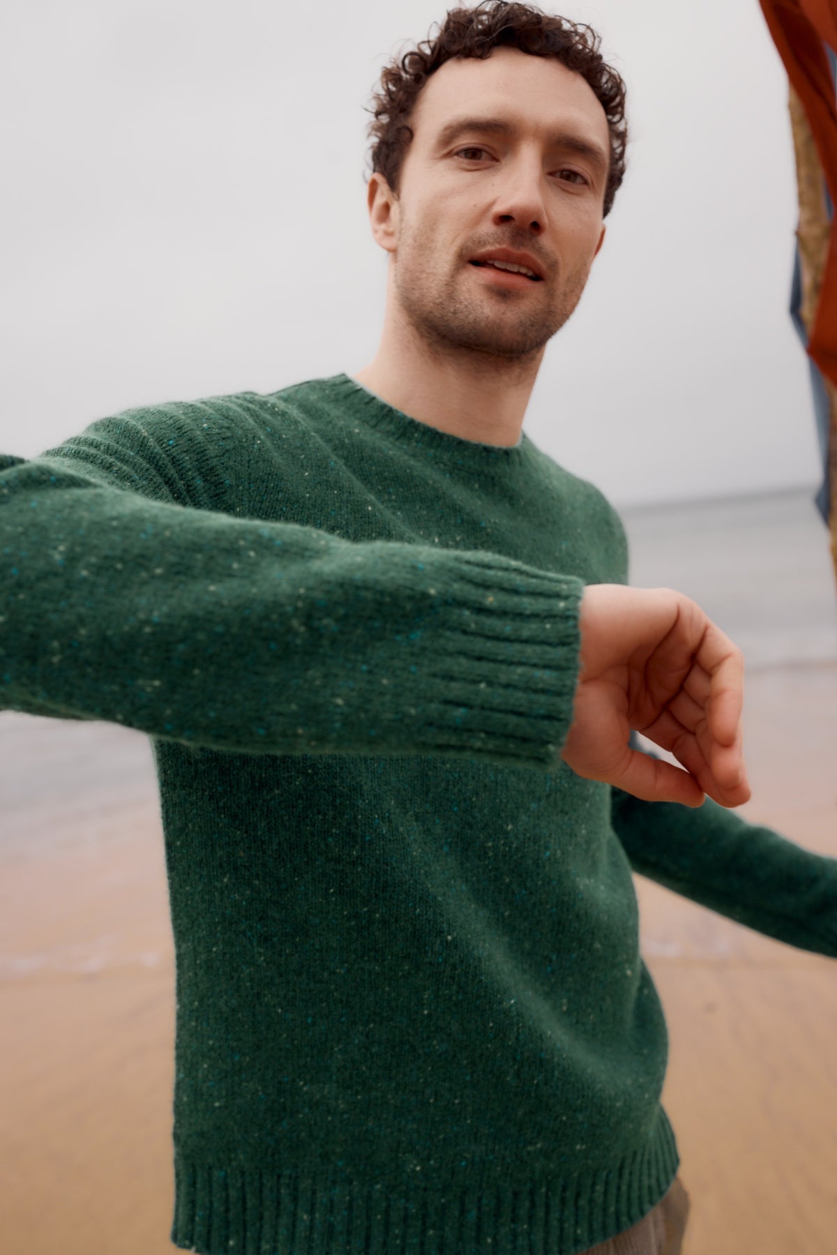 A man wearing a green Seasalt Cornwall merino wool sweater standing on a sandy beach.