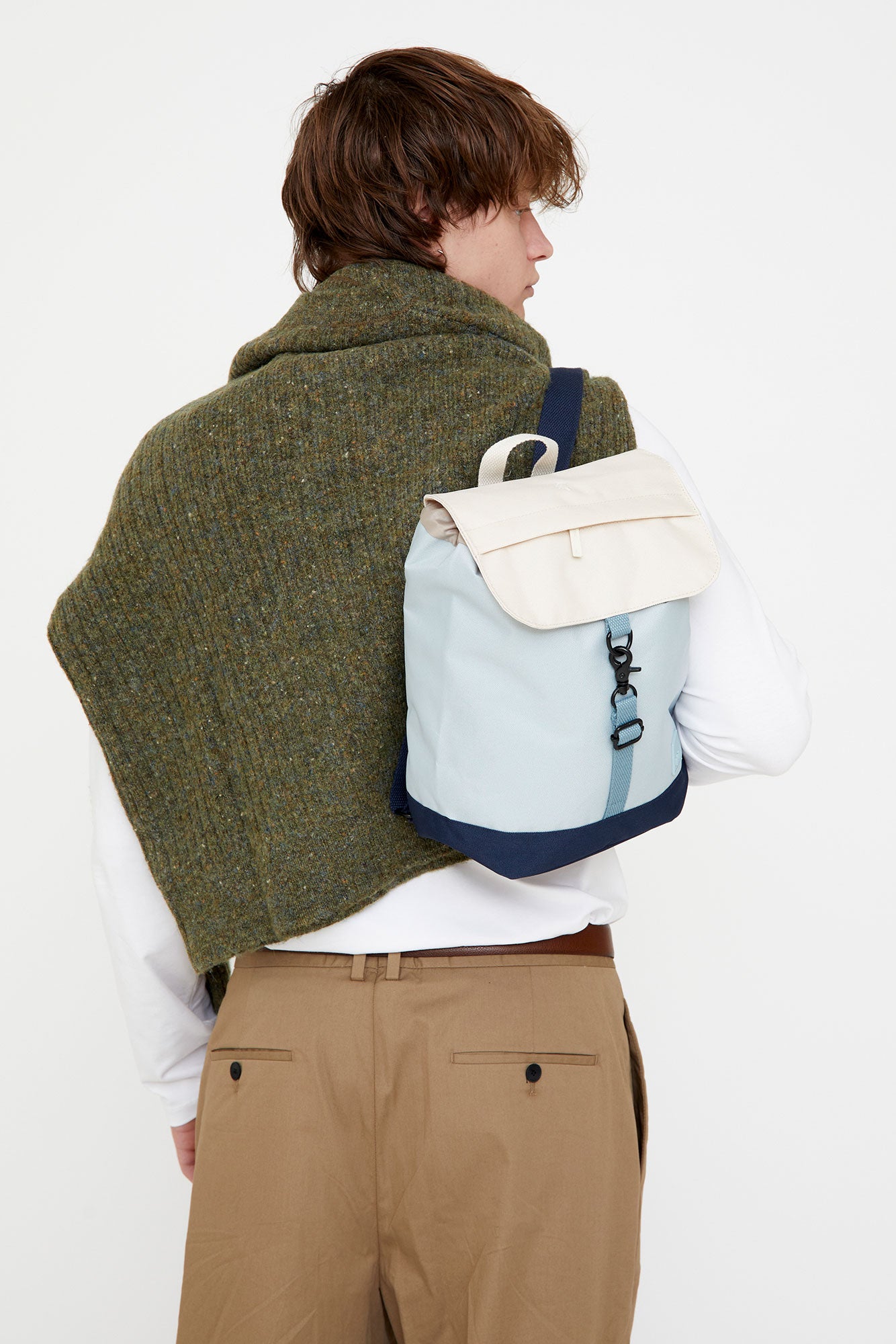 Person wearing a green coat, white shirt, beige pants, and carrying a light blue backpack with a white bag on a white background.