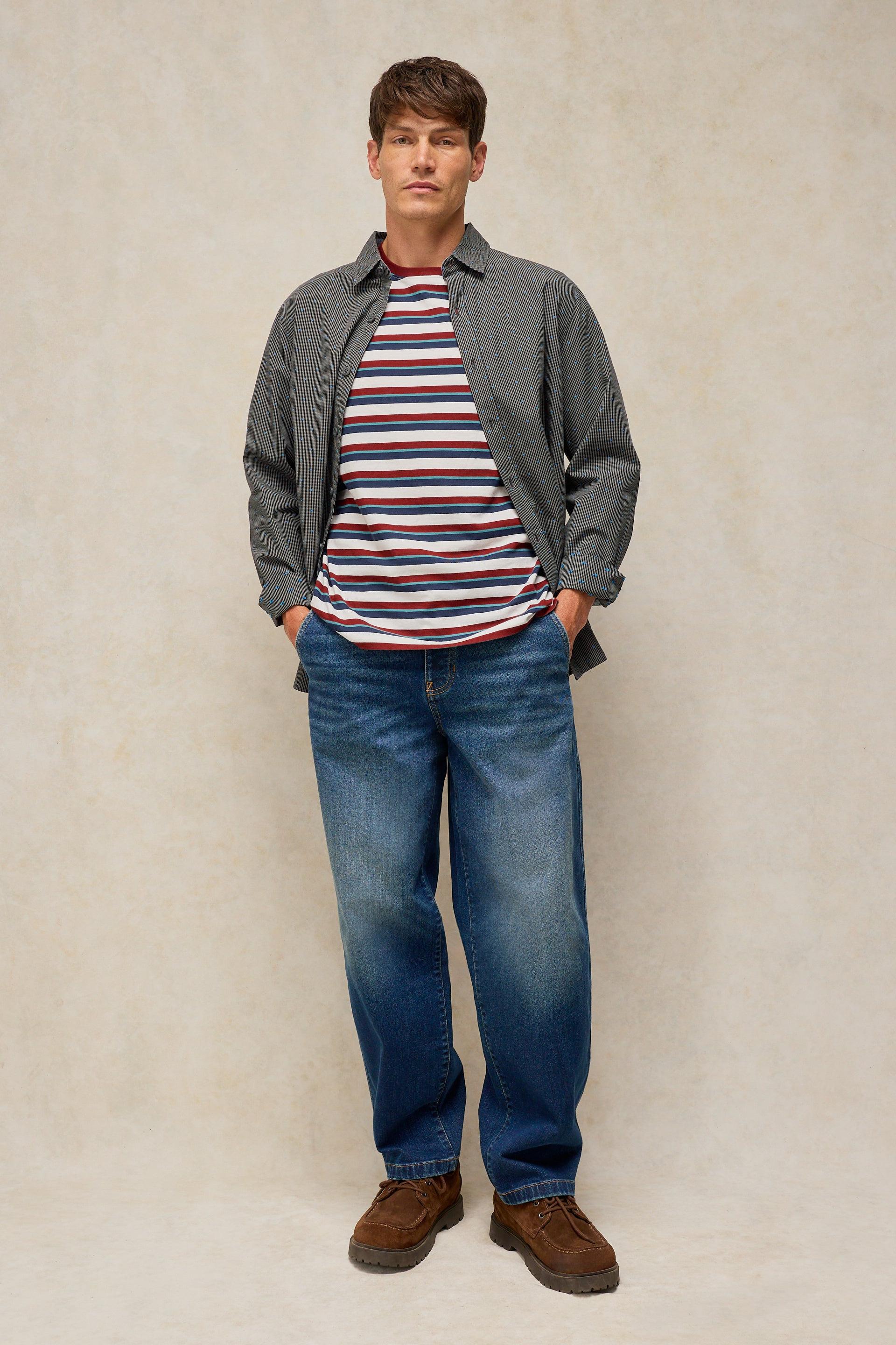 A man modeling a casual outfit consisting of a dark gray button-up overshirt, a red, white, and navy blue striped t-shirt, blue jeans, and brown suede boots, standing against a textured beige studio backdrop.