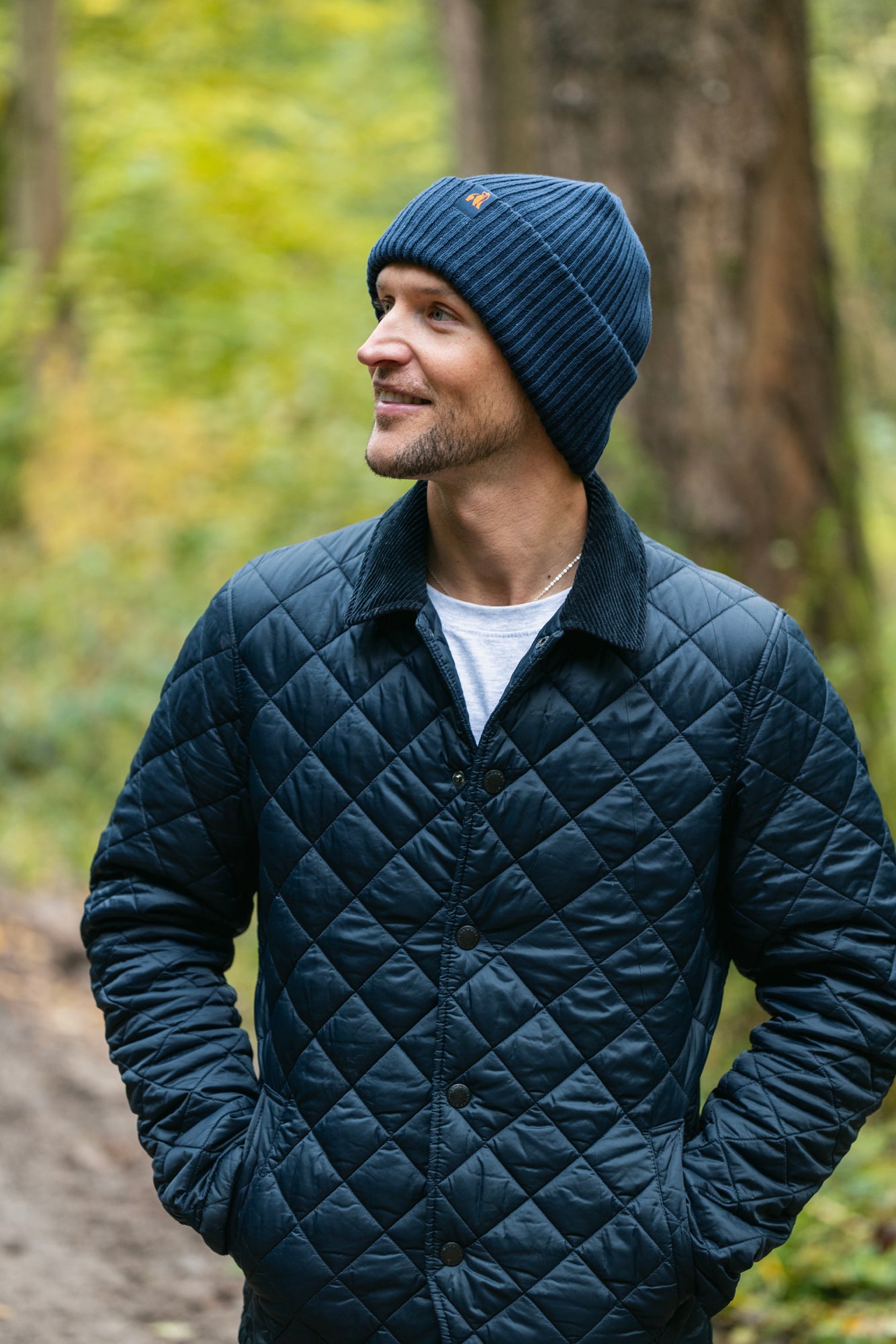 A man wearing a navy blue Swole Panda beanie and a quilted jacket standing in a forest.