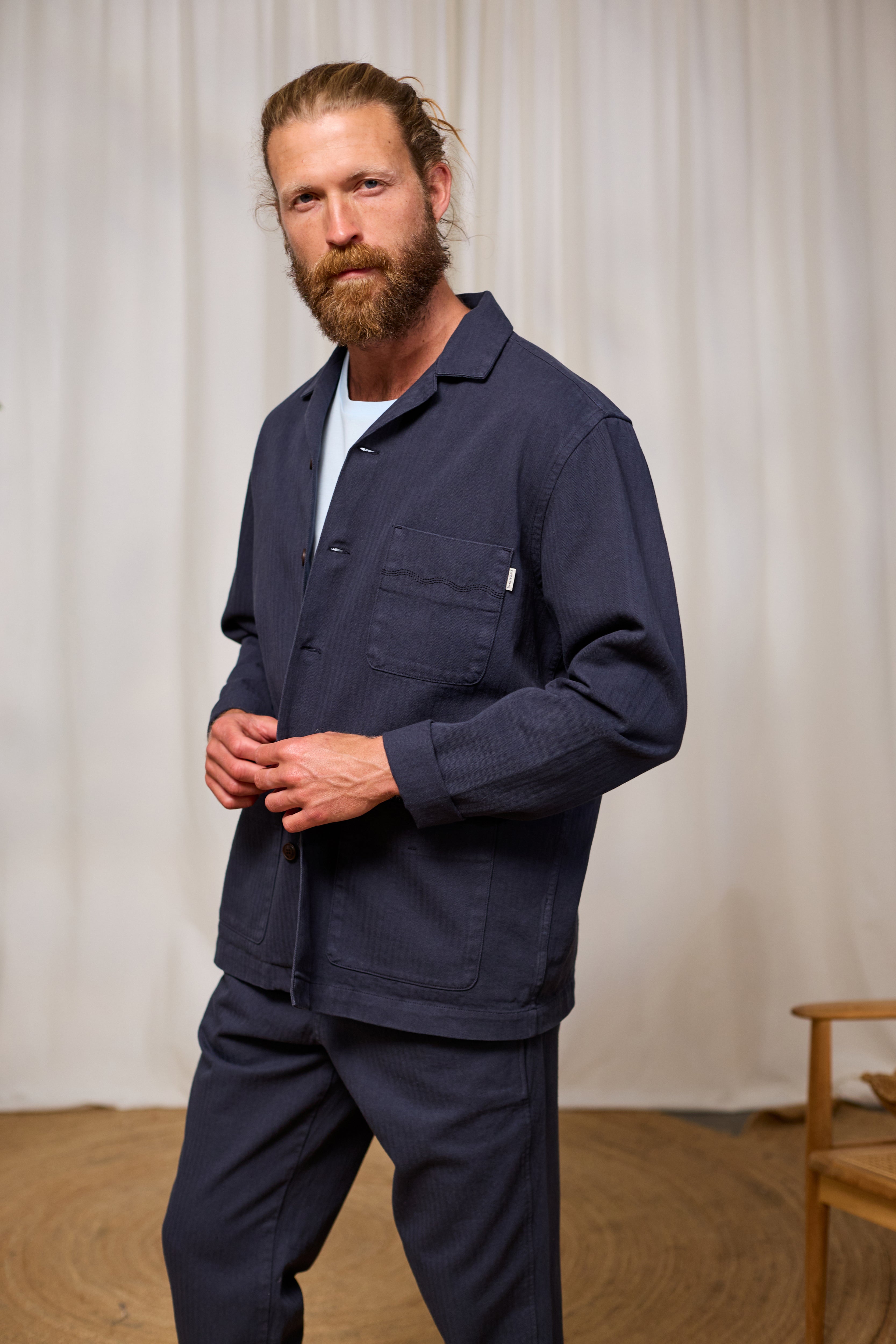 Man wearing a navy blue jacket and pants standing against a white curtain.